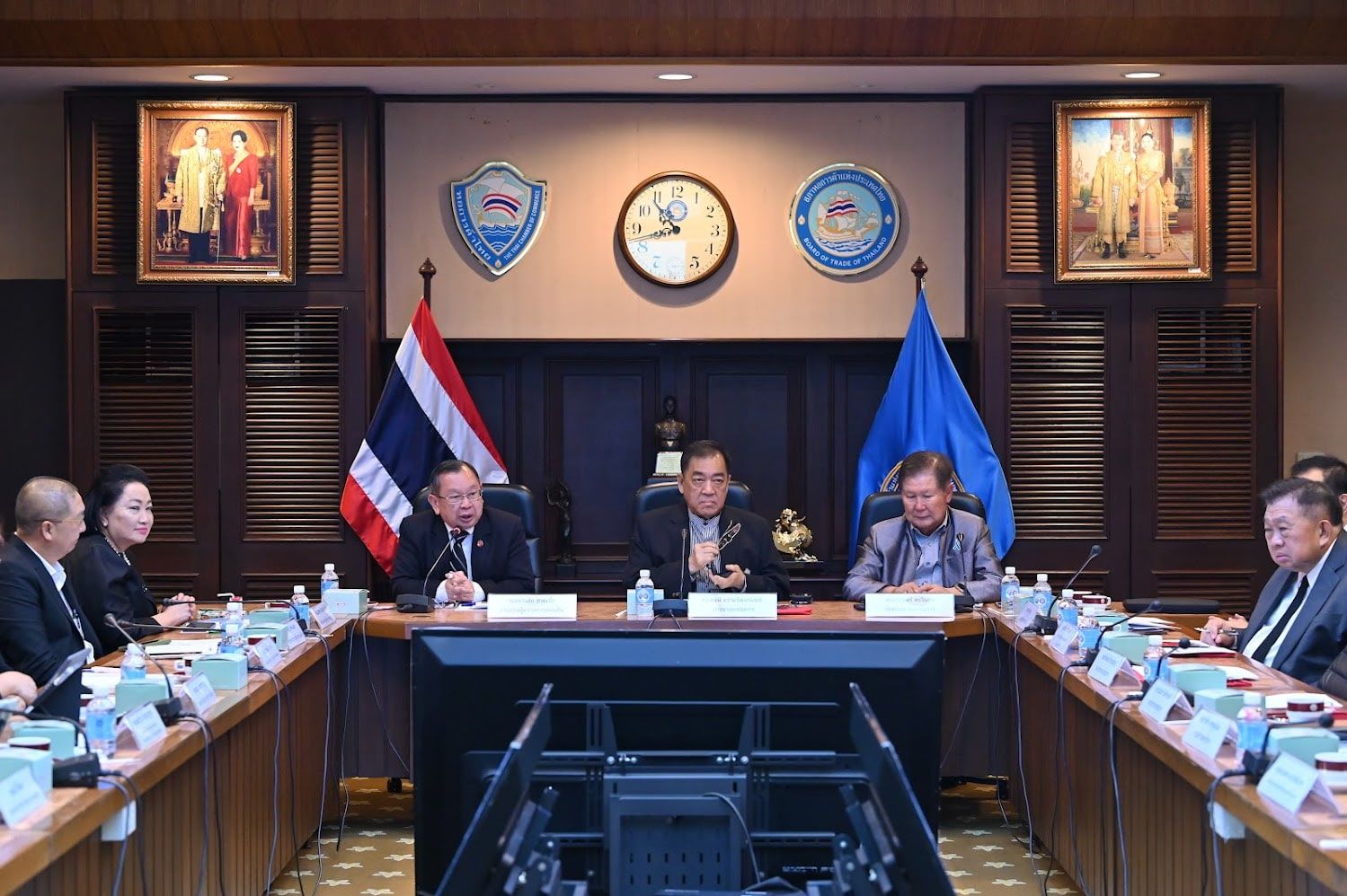 Group of officials in a formal meeting room with a Thai flag and portraits.
