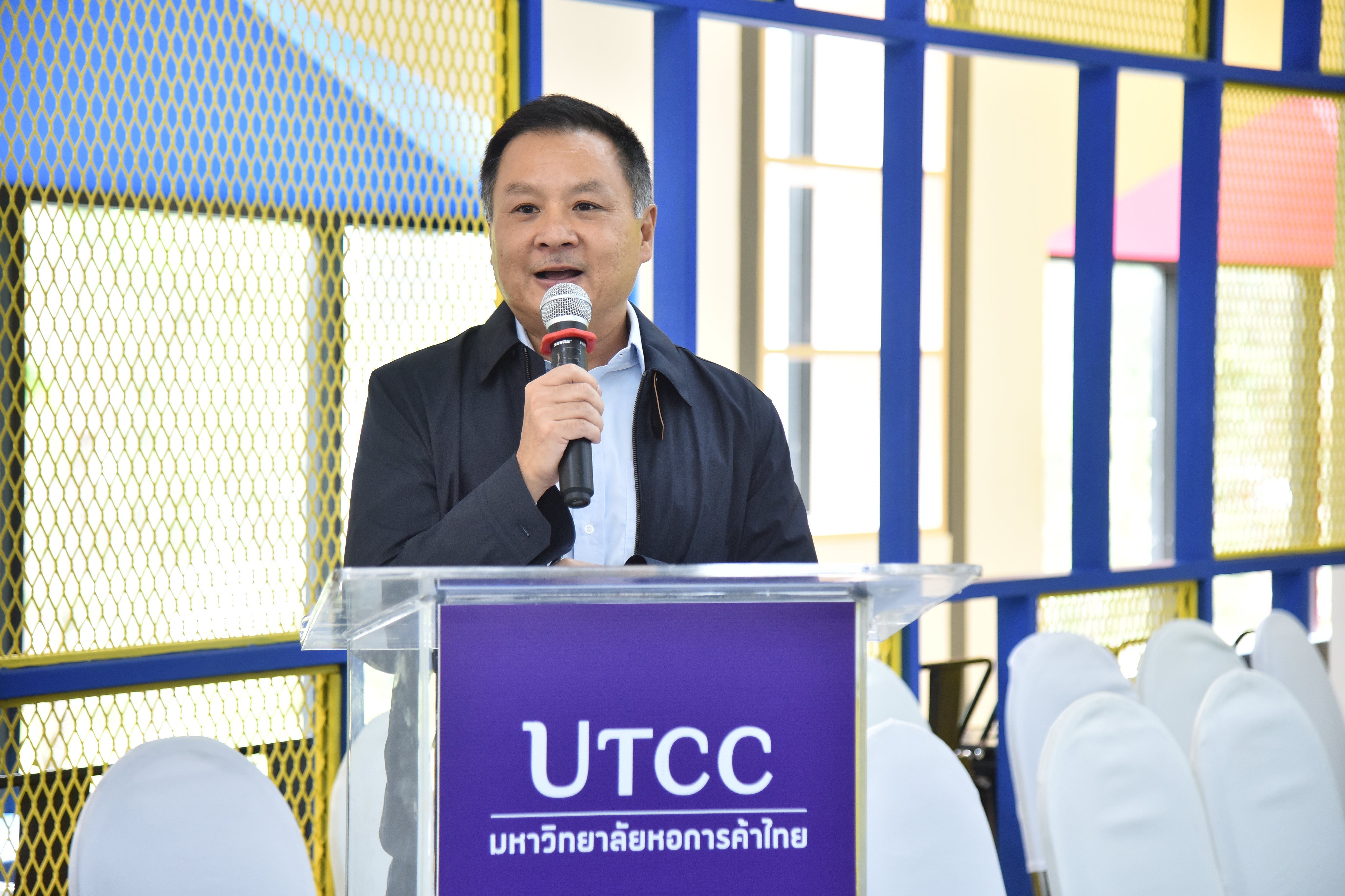 A man in a dark jacket speaks into a microphone at a UTCC podium.