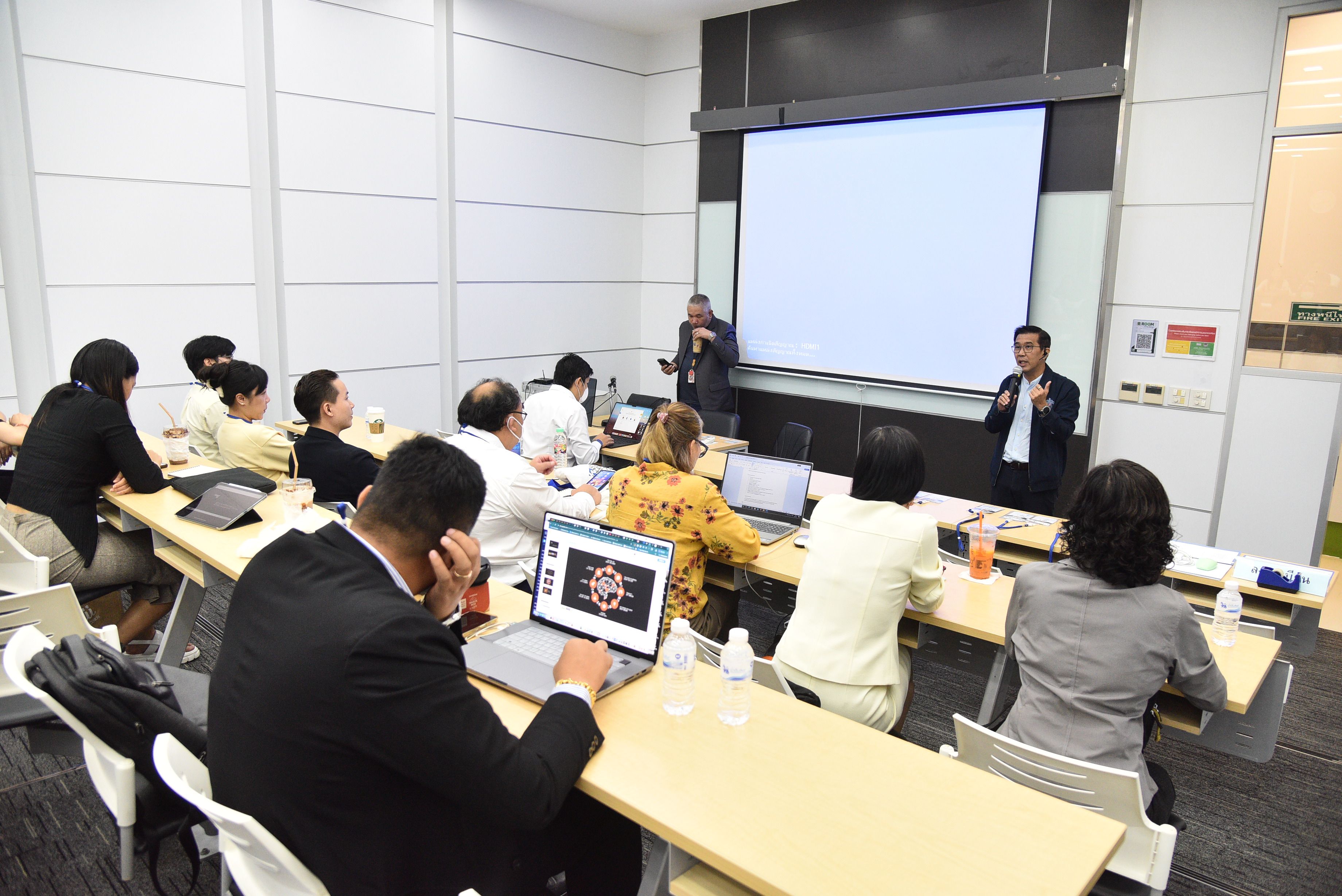 Attendees listen to speakers in a modern classroom with a projection screen during a presentation.