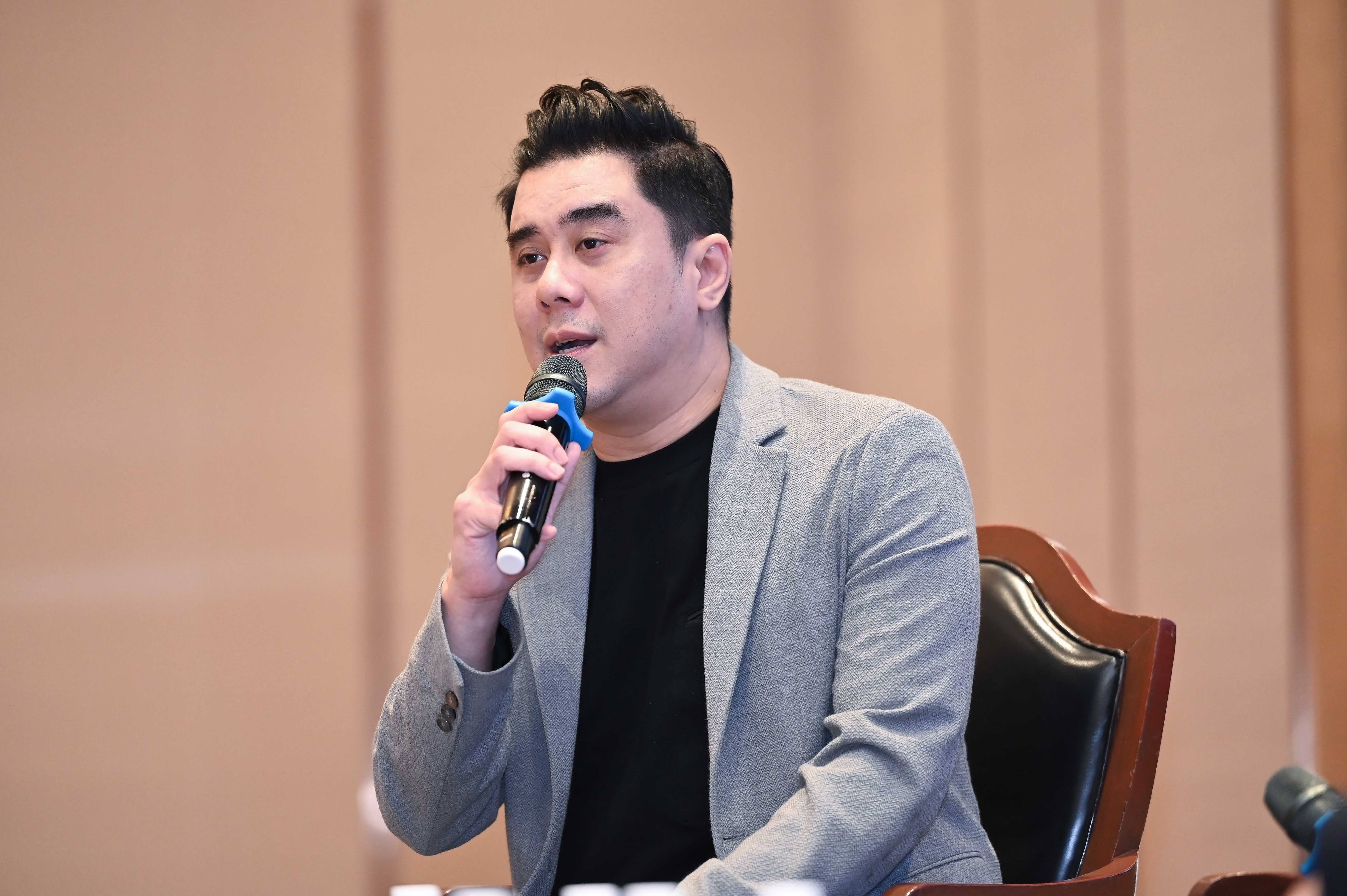 A man in a grey blazer speaks into a microphone at a professional event.