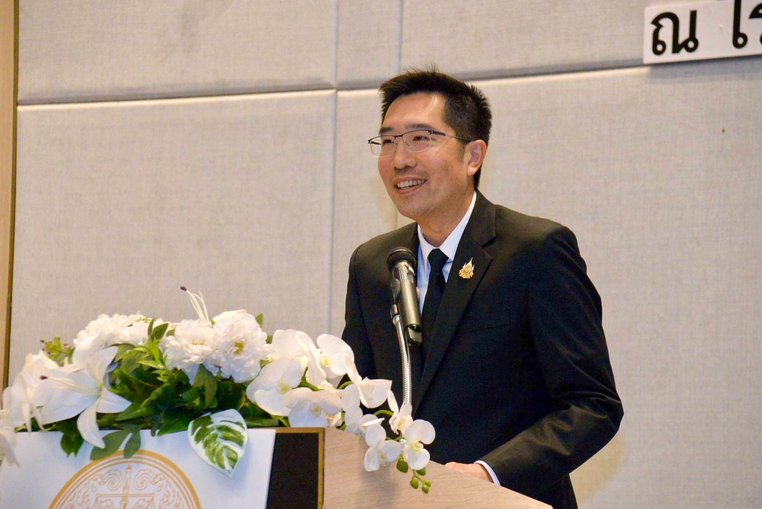 A smiling man in a suit and glasses speaks at a podium adorned with white flowers.