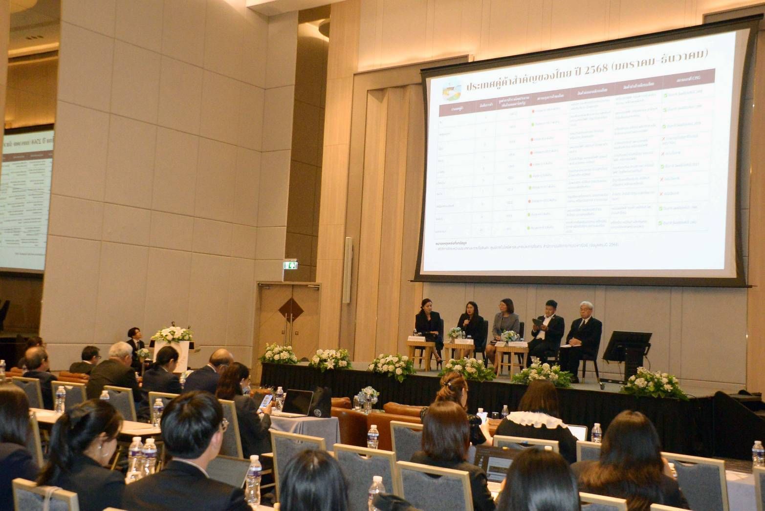 A panel of five speakers presents to an audience in a large conference room.