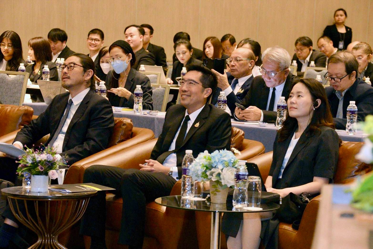 Professionals in dark suits attending a conference, seated in a formal audience setting.