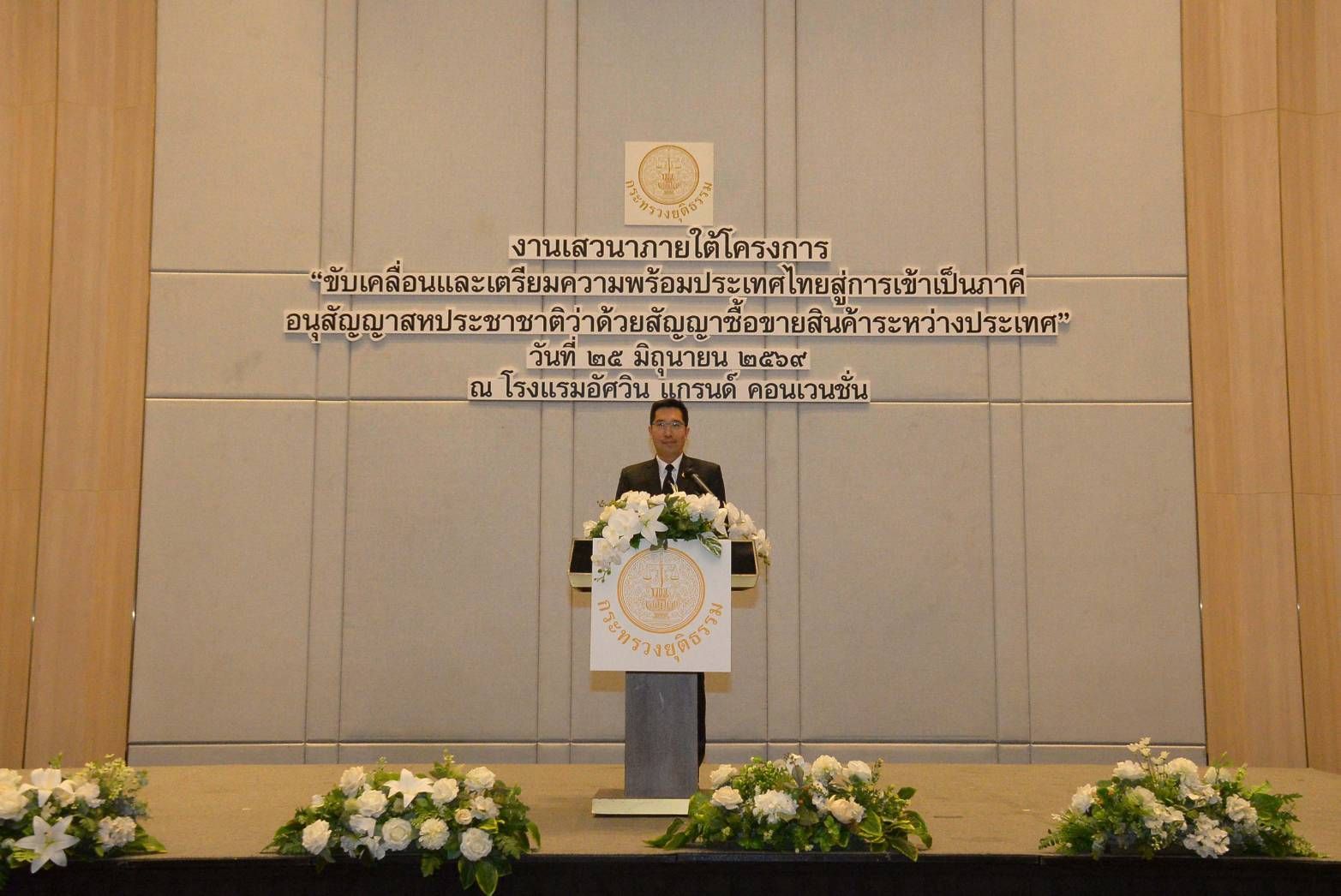 A man in a suit speaks at a podium with white flowers and Thai text on a wall.