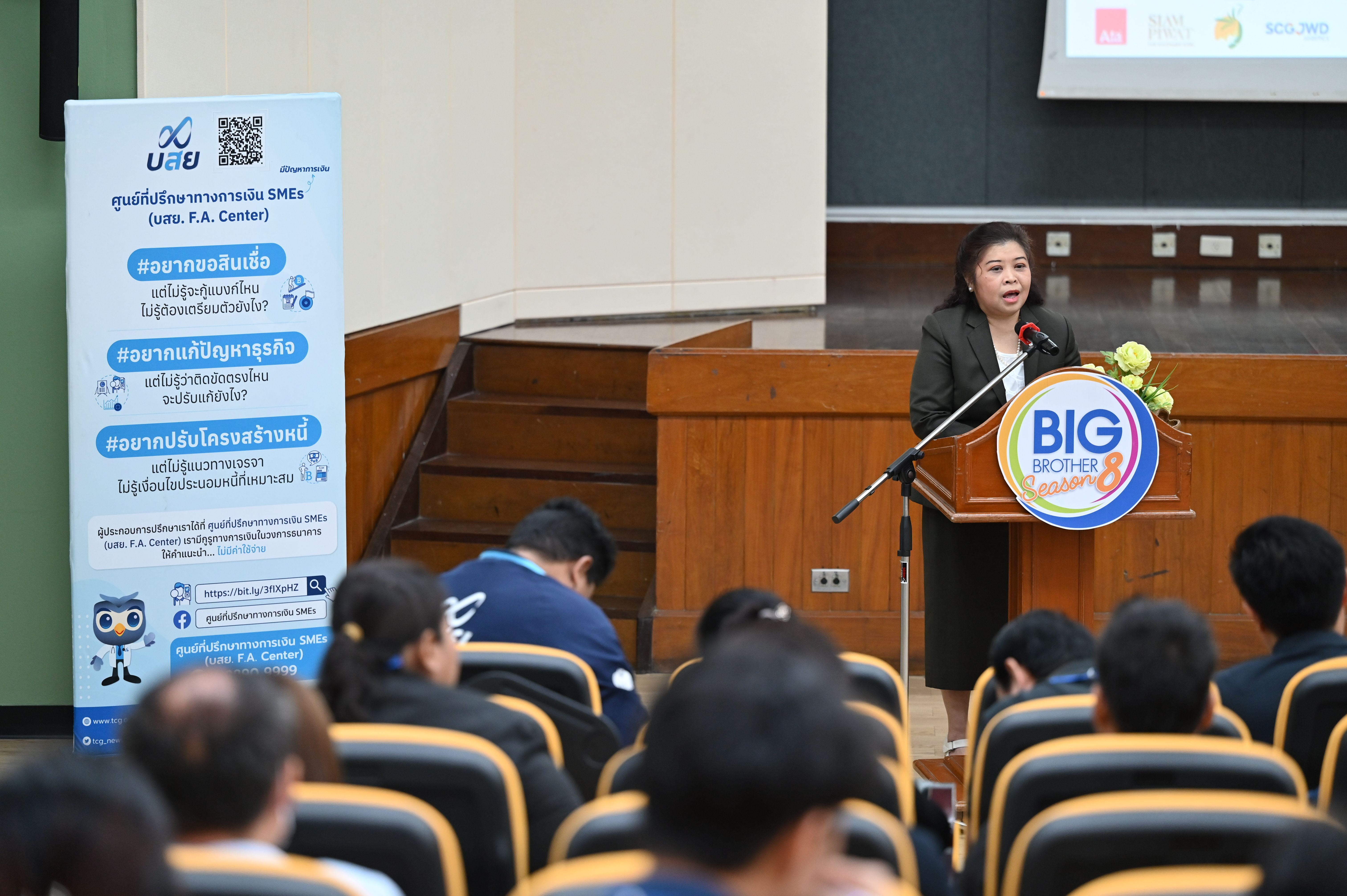 Woman speaks at a podium to an audience in a lecture hall with a banner.