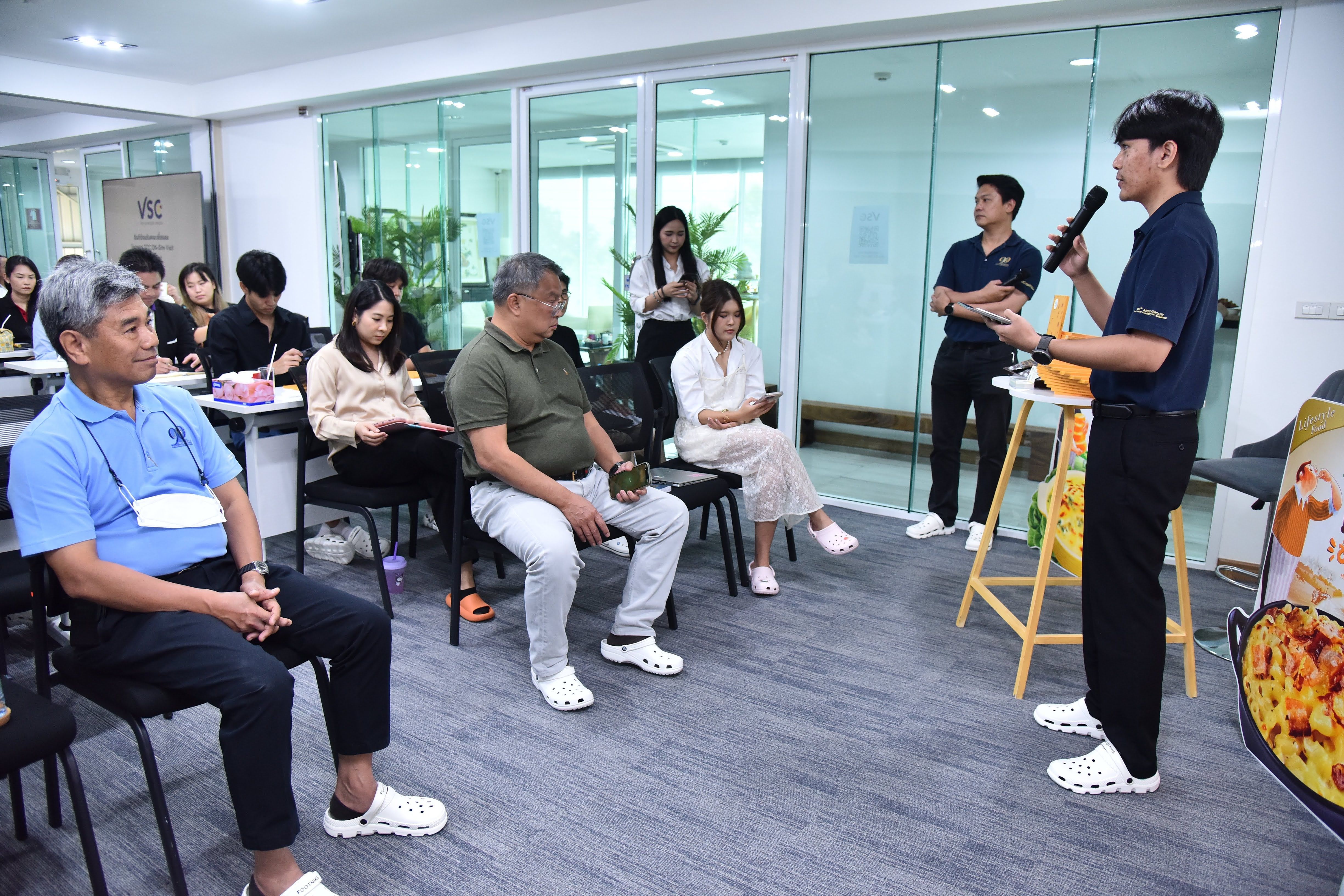 A speaker presents to a diverse, seated audience in a bright, modern classroom setting.