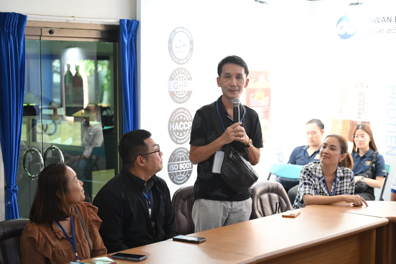 Man speaking into microphone addresses an audience during an indoor meeting.