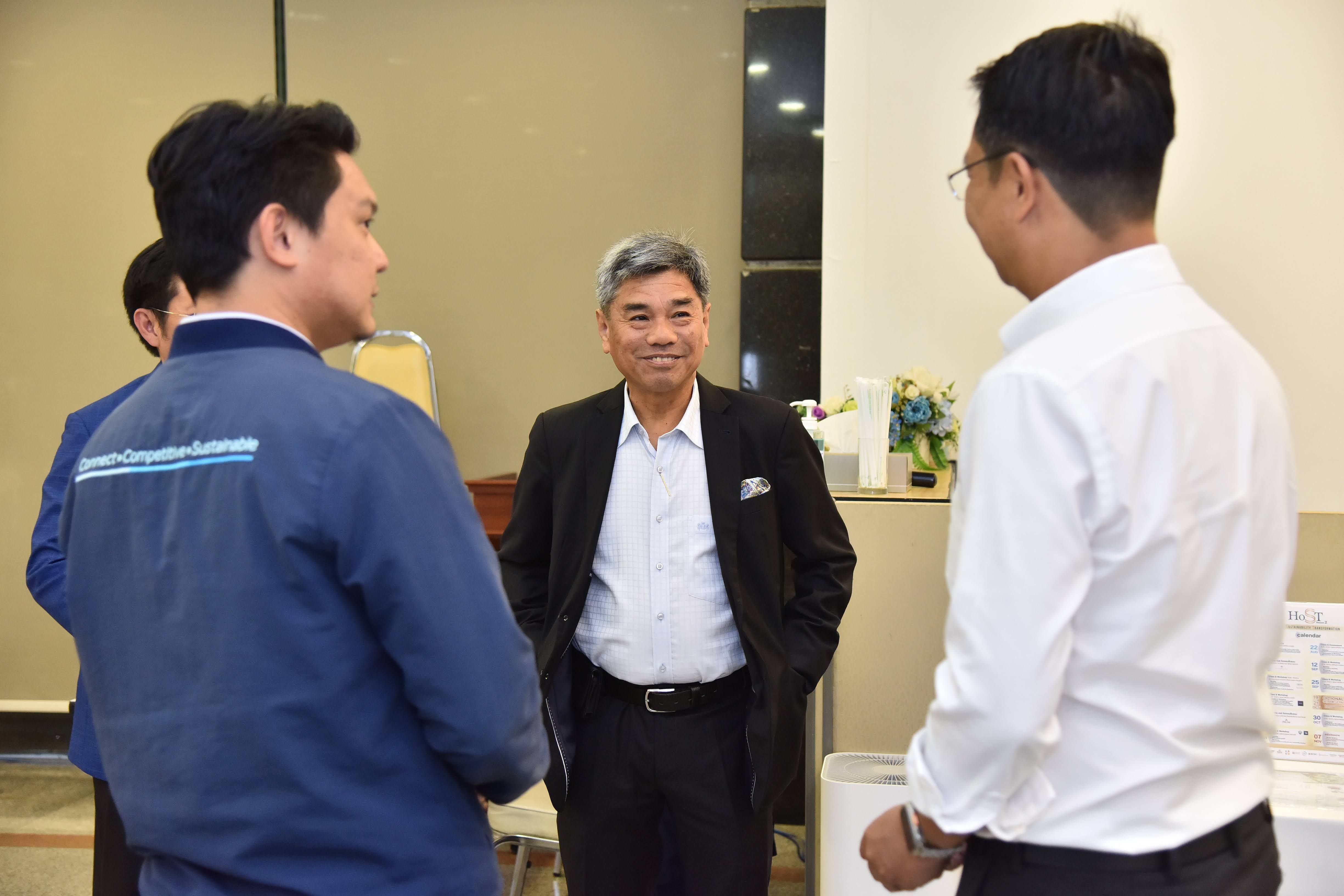 Three men converse, an older man in a suit smiles at a younger colleague.