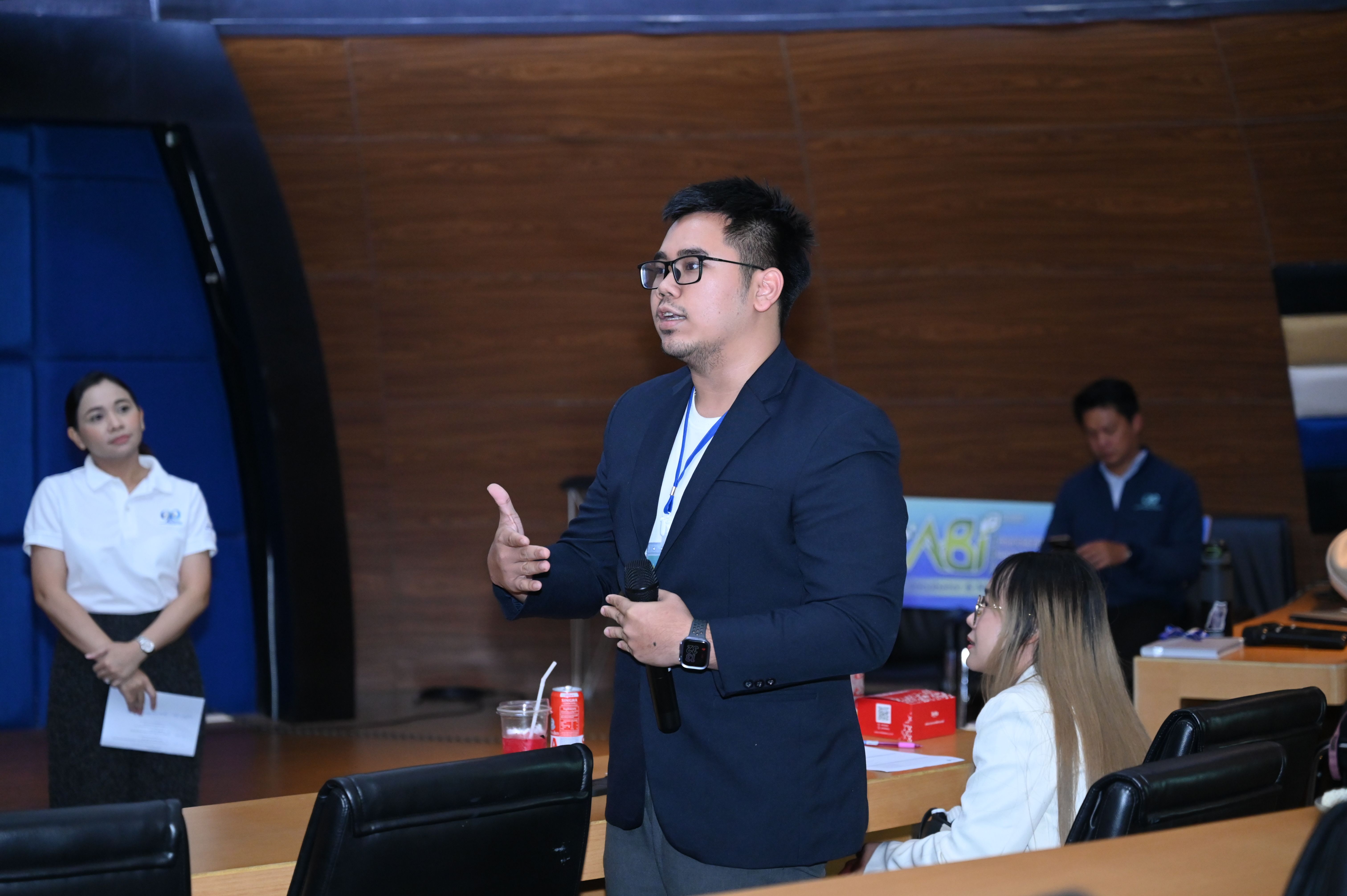 A professional man in a blazer and glasses speaks, gesturing during a presentation.