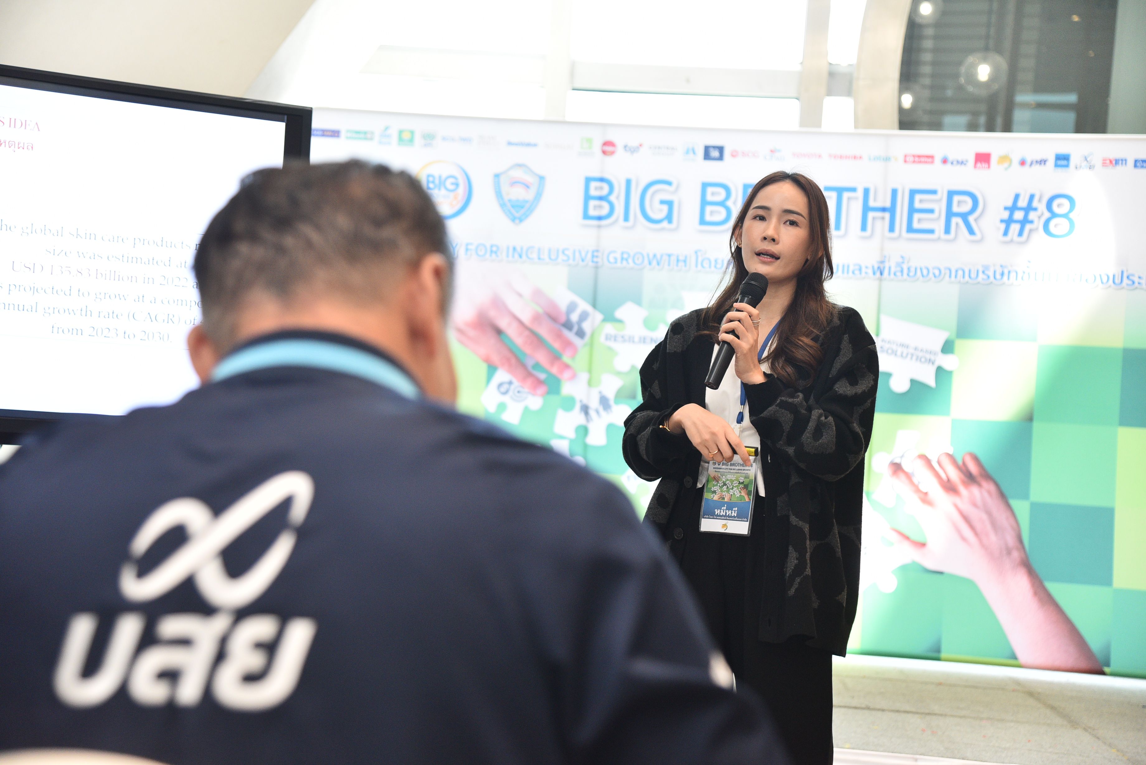 A woman speaks into a microphone at a conference, with a 'BIG BROTHER #8' banner.