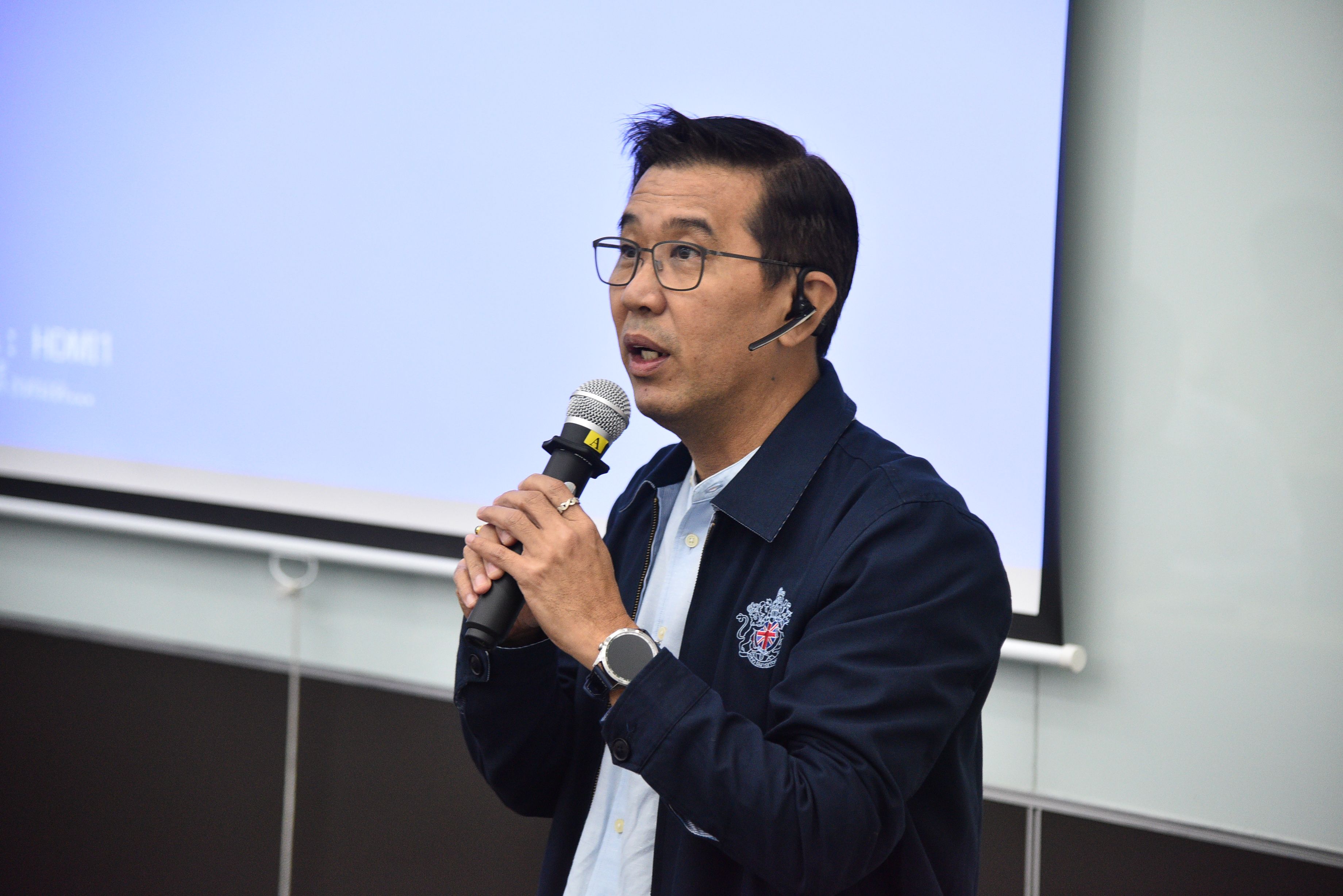 A man wearing glasses and a dark jacket speaks into a microphone.