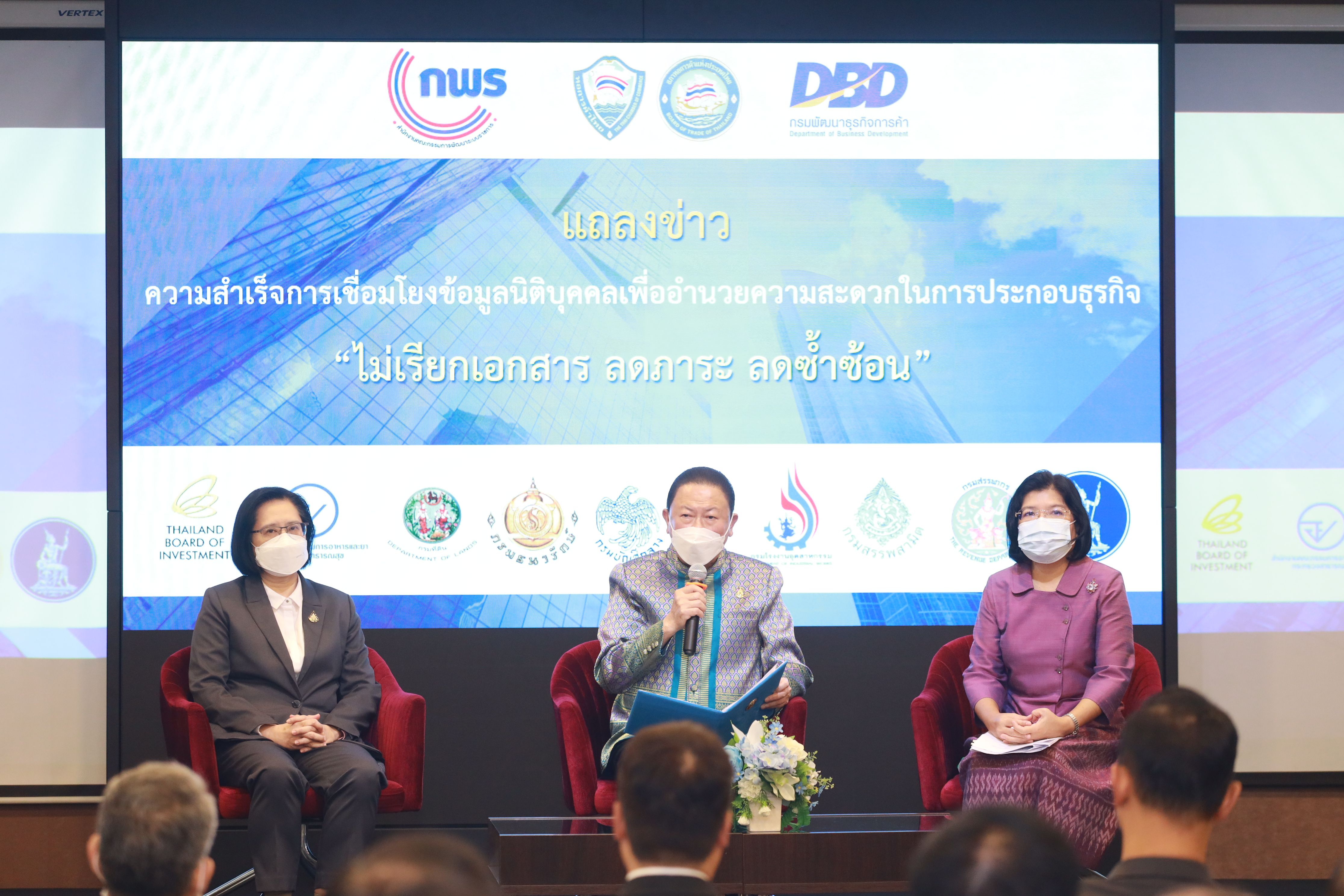 Officials on stage, one speaking into a microphone, with a large screen displaying logos.