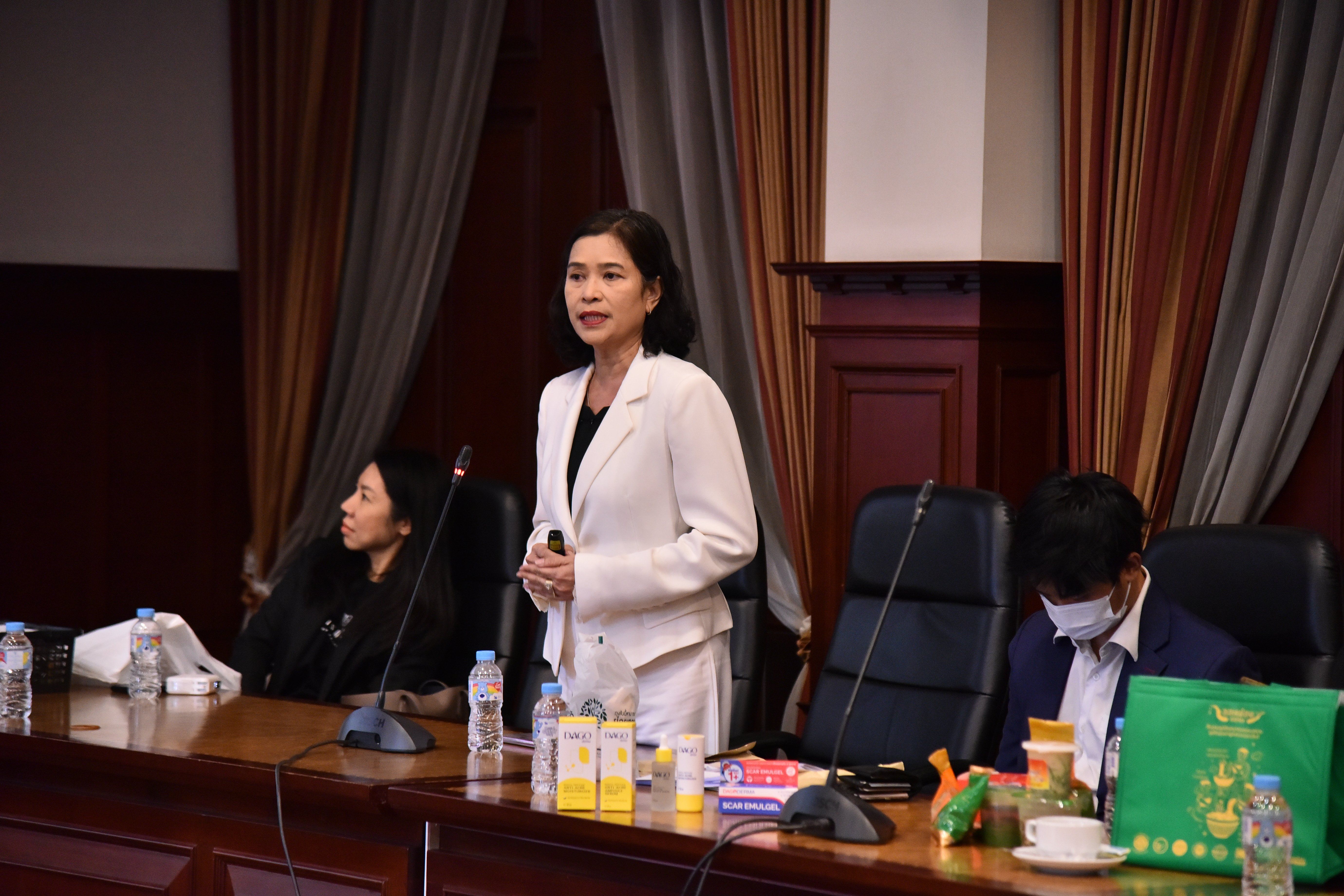 Woman in white blazer speaks at a formal conference table with other attendees.