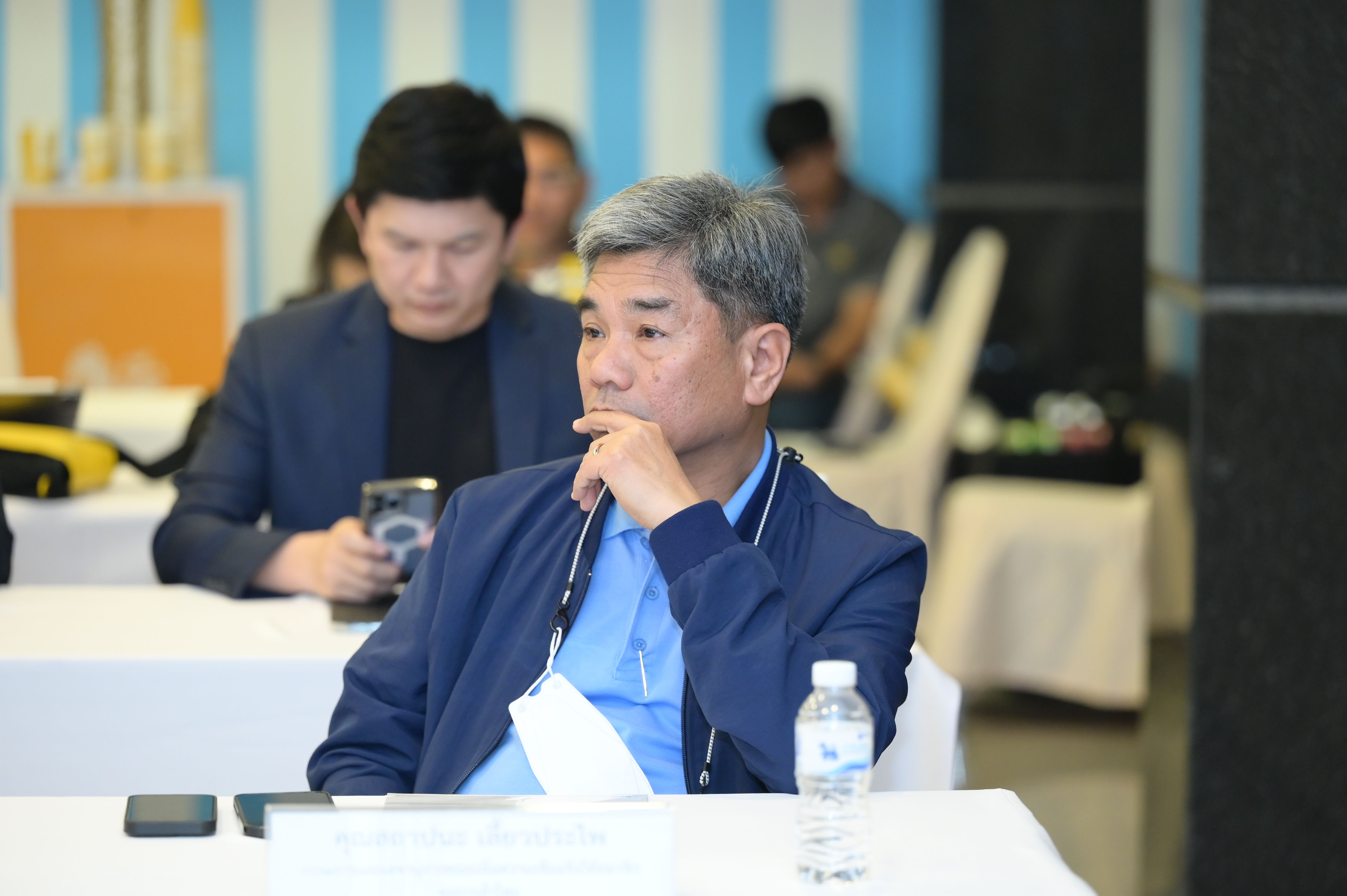 Middle-aged Asian man in a blue jacket sits thoughtfully at a conference table.