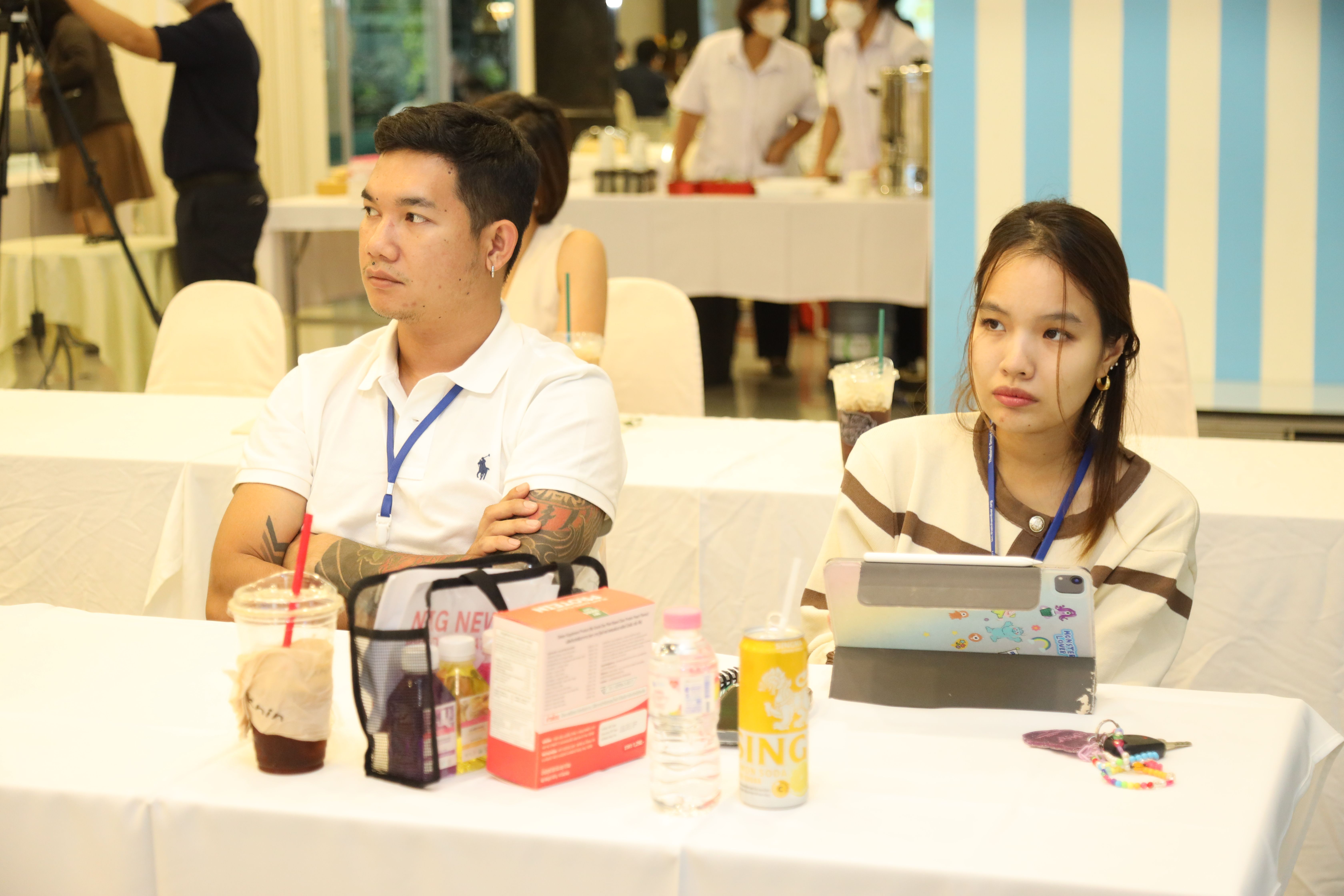 Man and woman with lanyards sit at a table, attentively observing an indoor event.