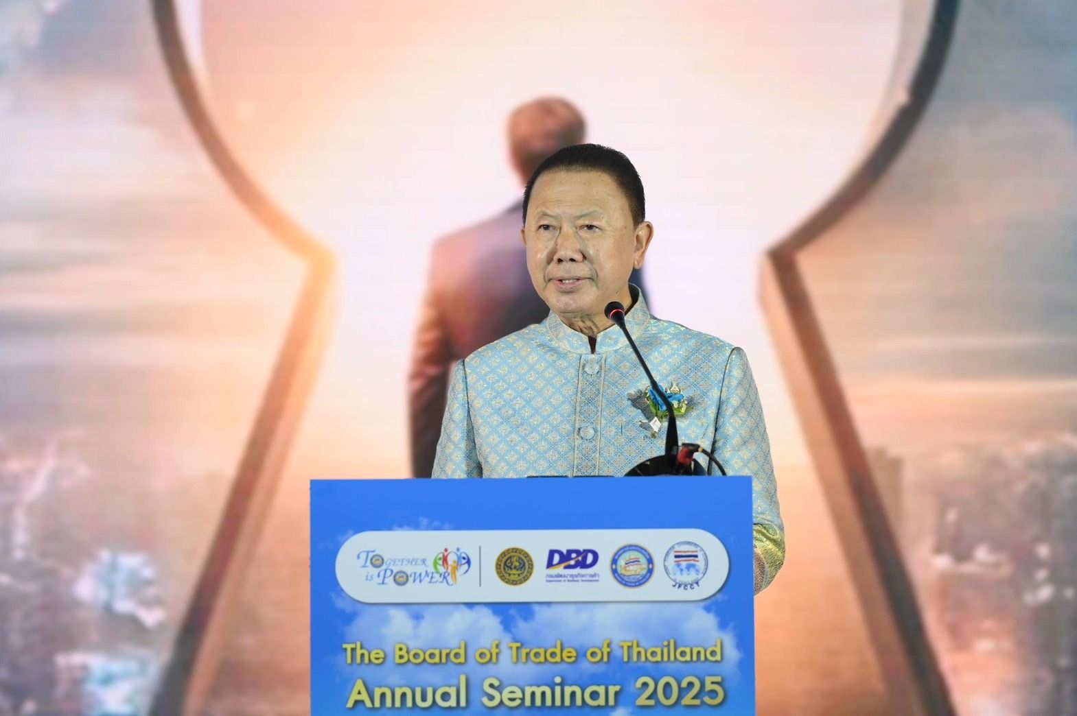 Man in traditional attire speaks at a podium with 'Annual Seminar 2025' sign.