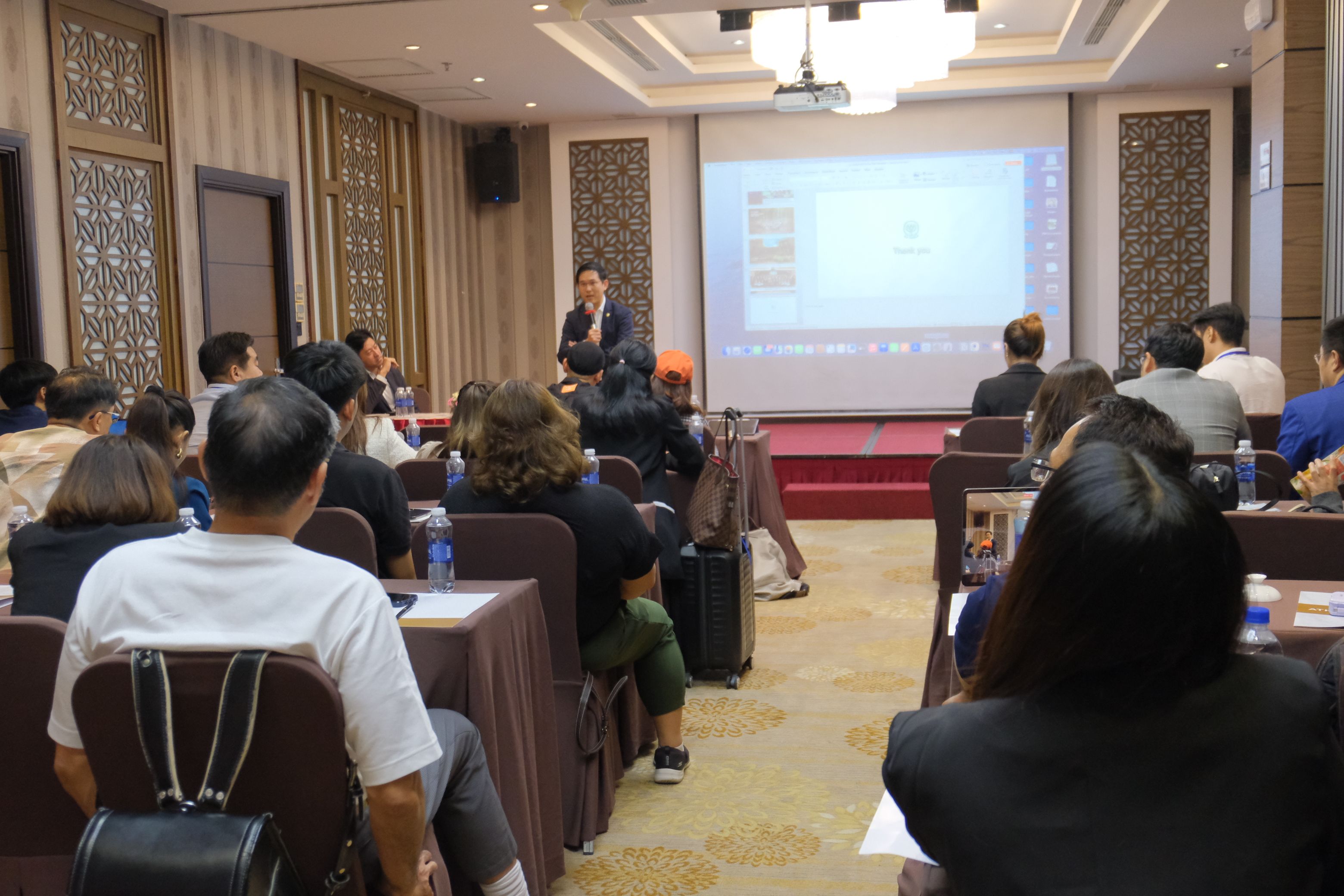 A speaker addresses an attentive audience in a well-lit conference room with a projector.