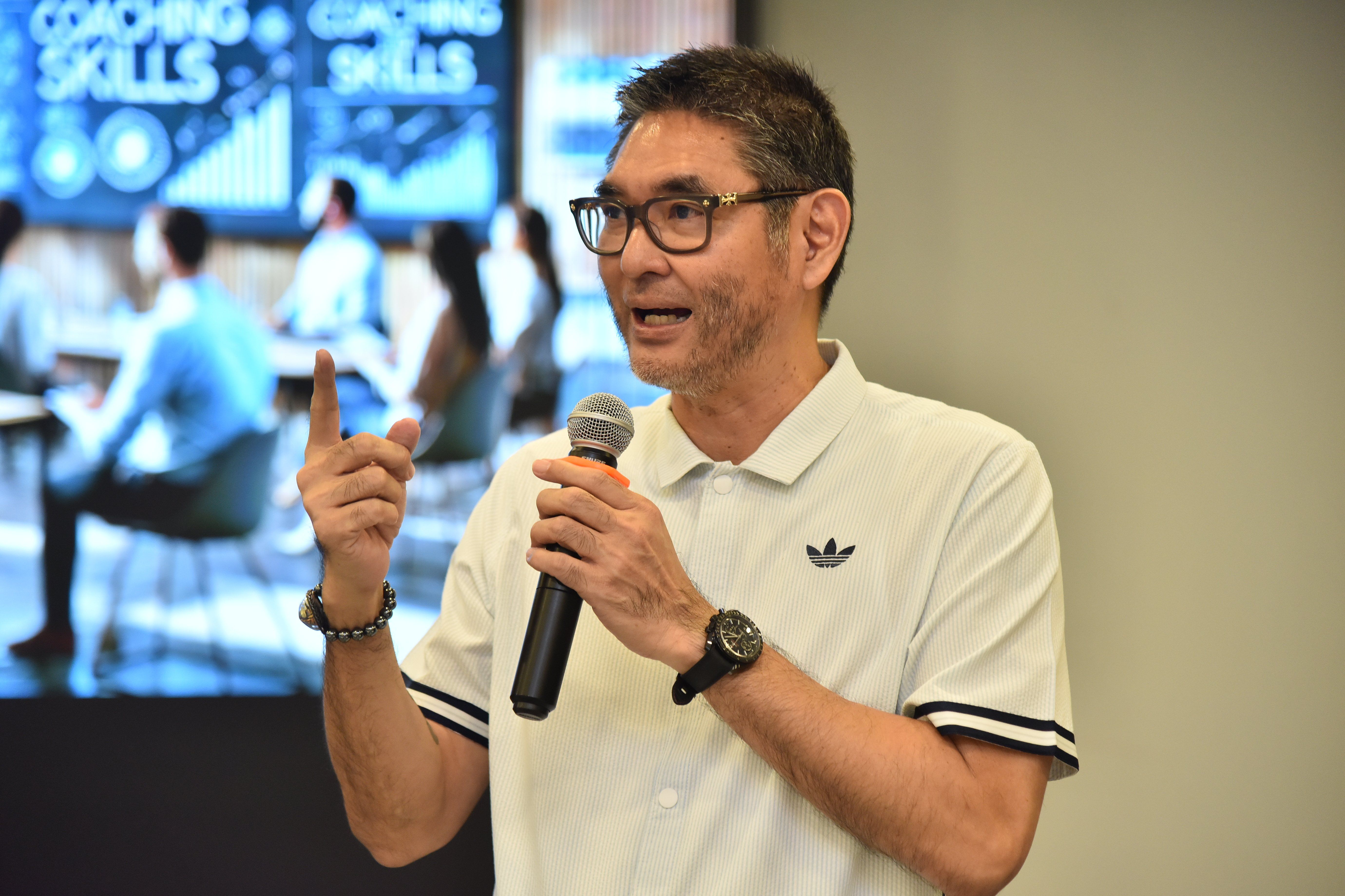 Man speaks into microphone wearing glasses and Adidas polo, gesturing with his hand.