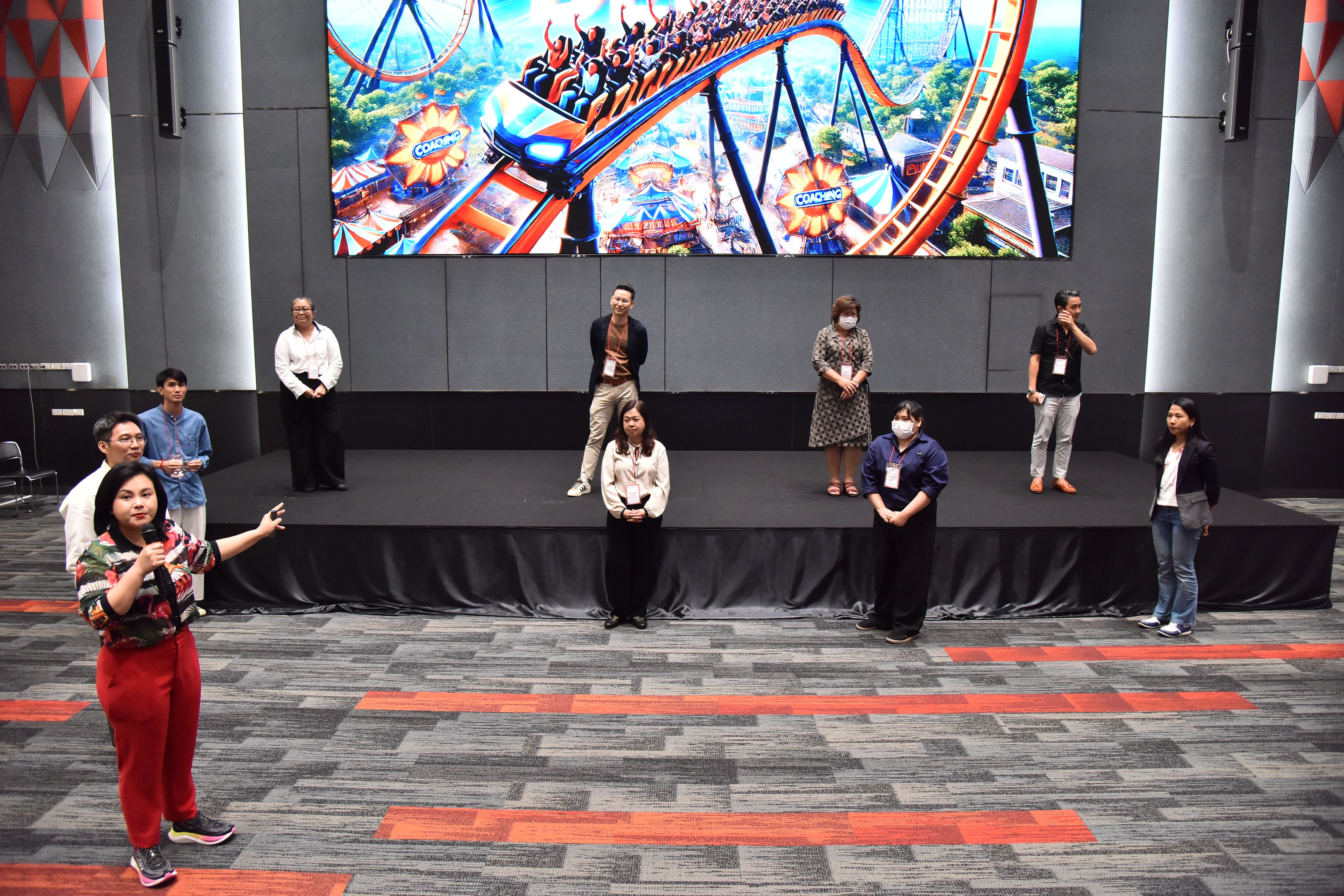 People gathered on a stage with a large screen displaying a vibrant roller coaster scene.