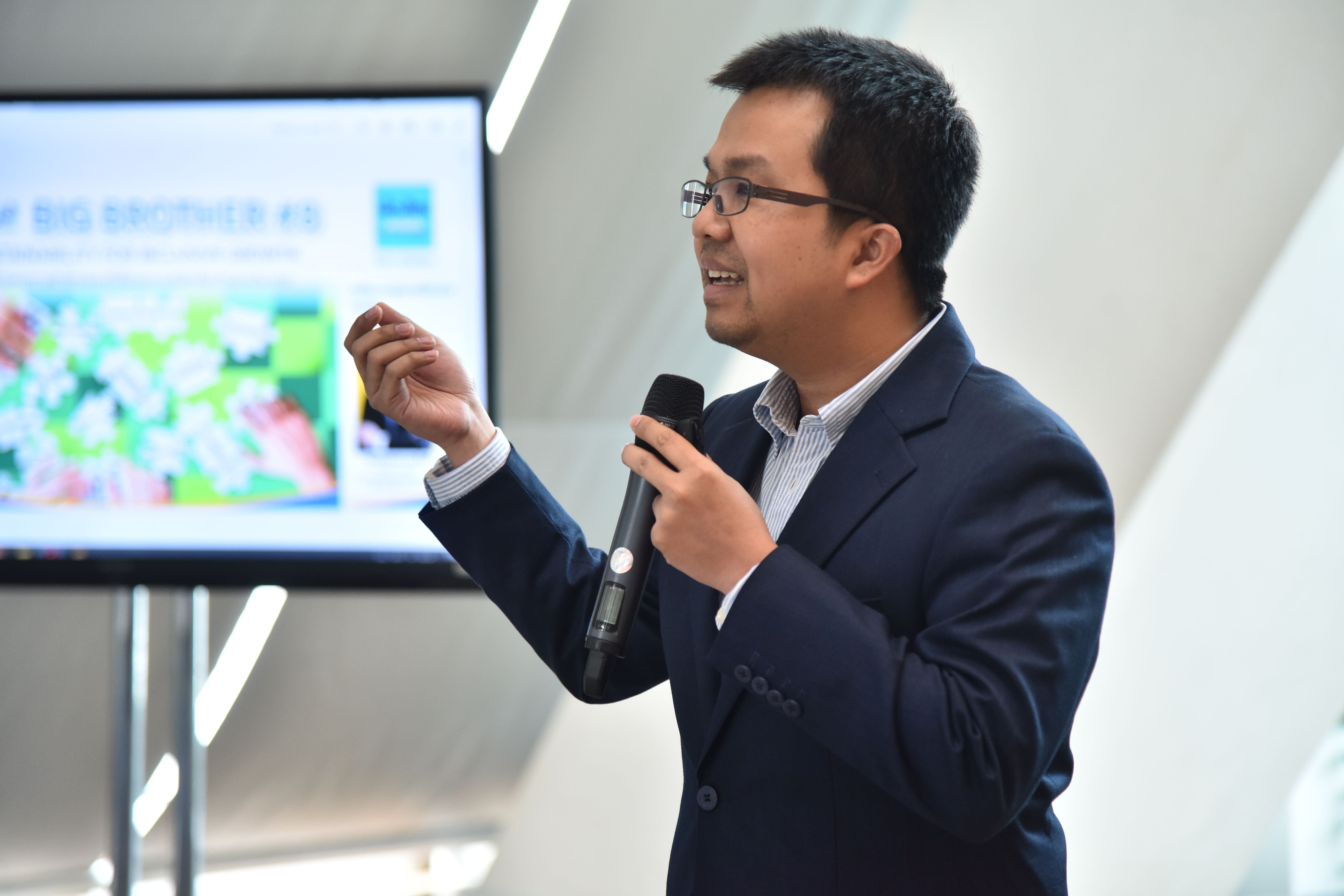 Businessman in glasses presenting with a microphone, screen visible in background.