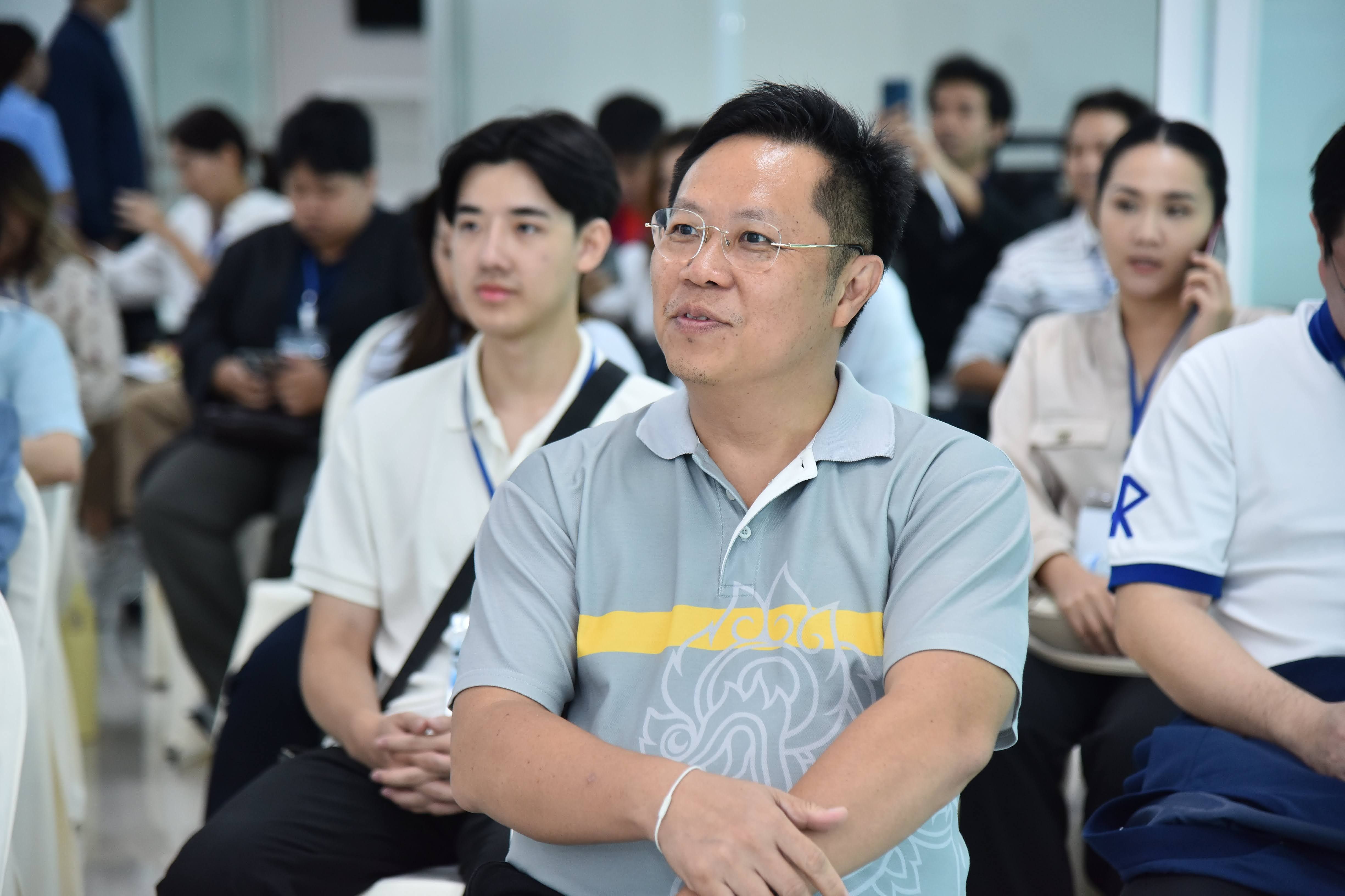 Asian man in glasses and grey polo shirt sits attentively, looking forward.