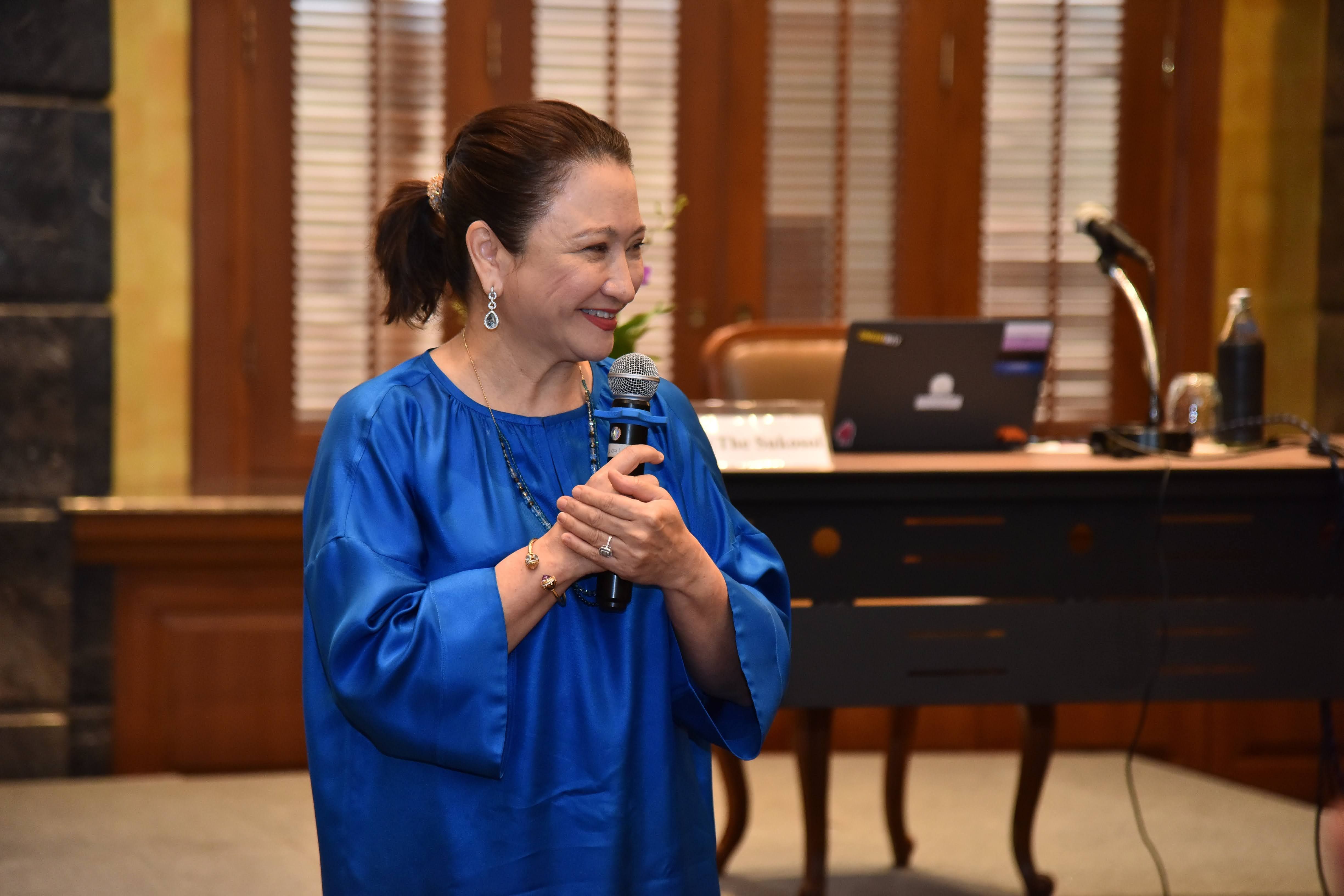 Smiling woman in a blue dress holds a microphone, speaking at an indoor event.