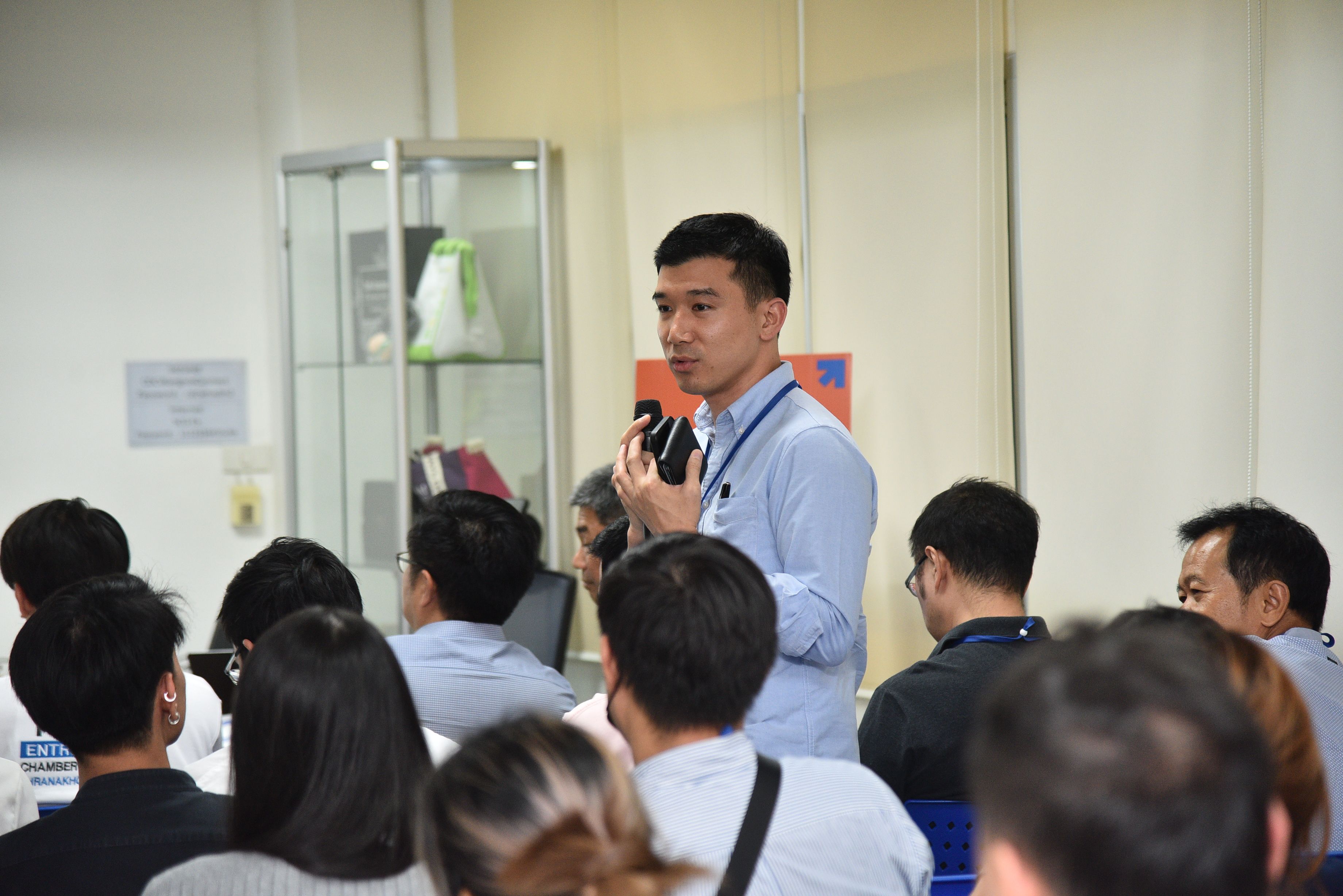 Man in light blue shirt speaks into a microphone to an audience in a bright room.