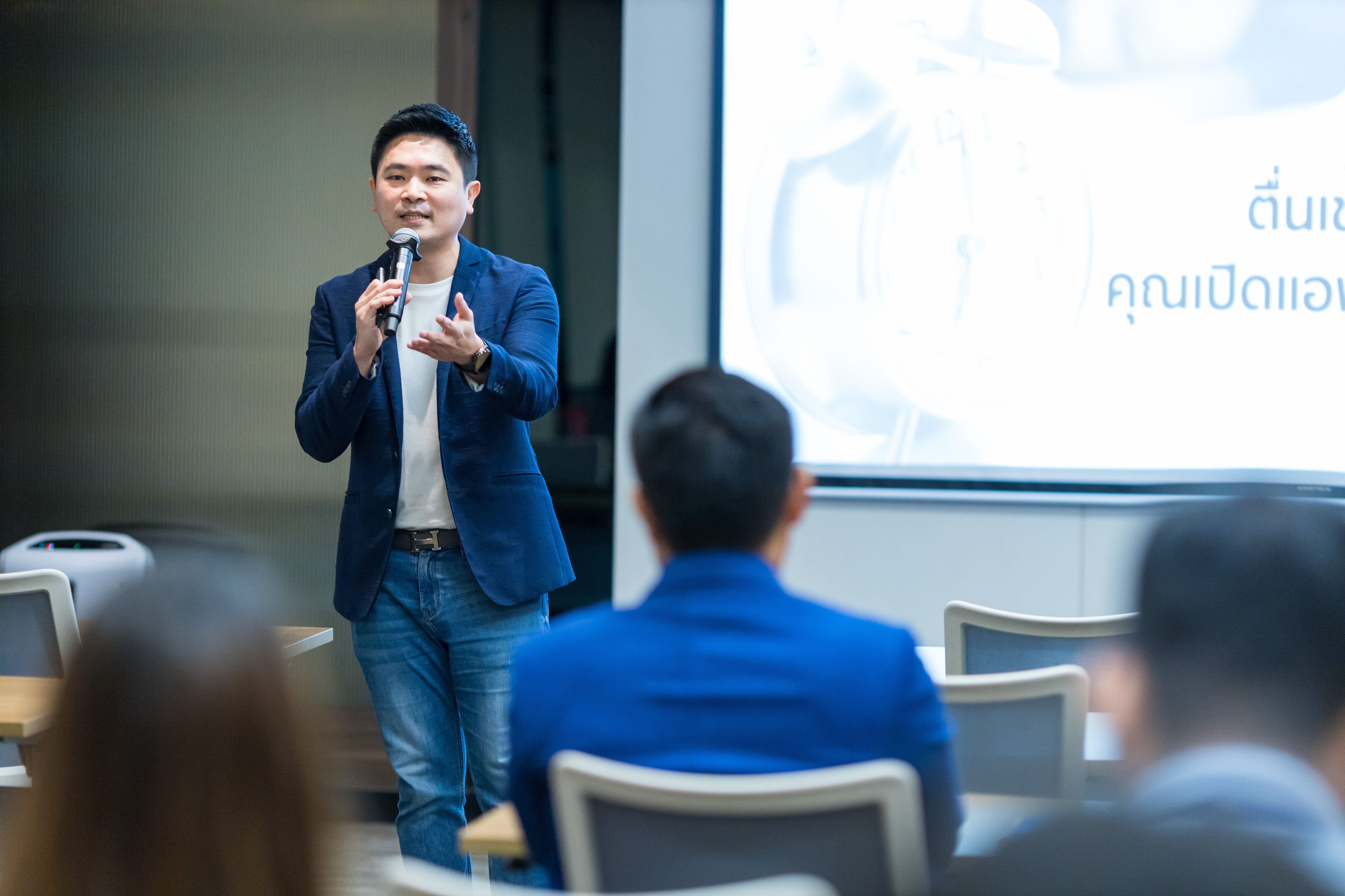 A man in a blue blazer speaks passionately into a microphone at a business presentation.