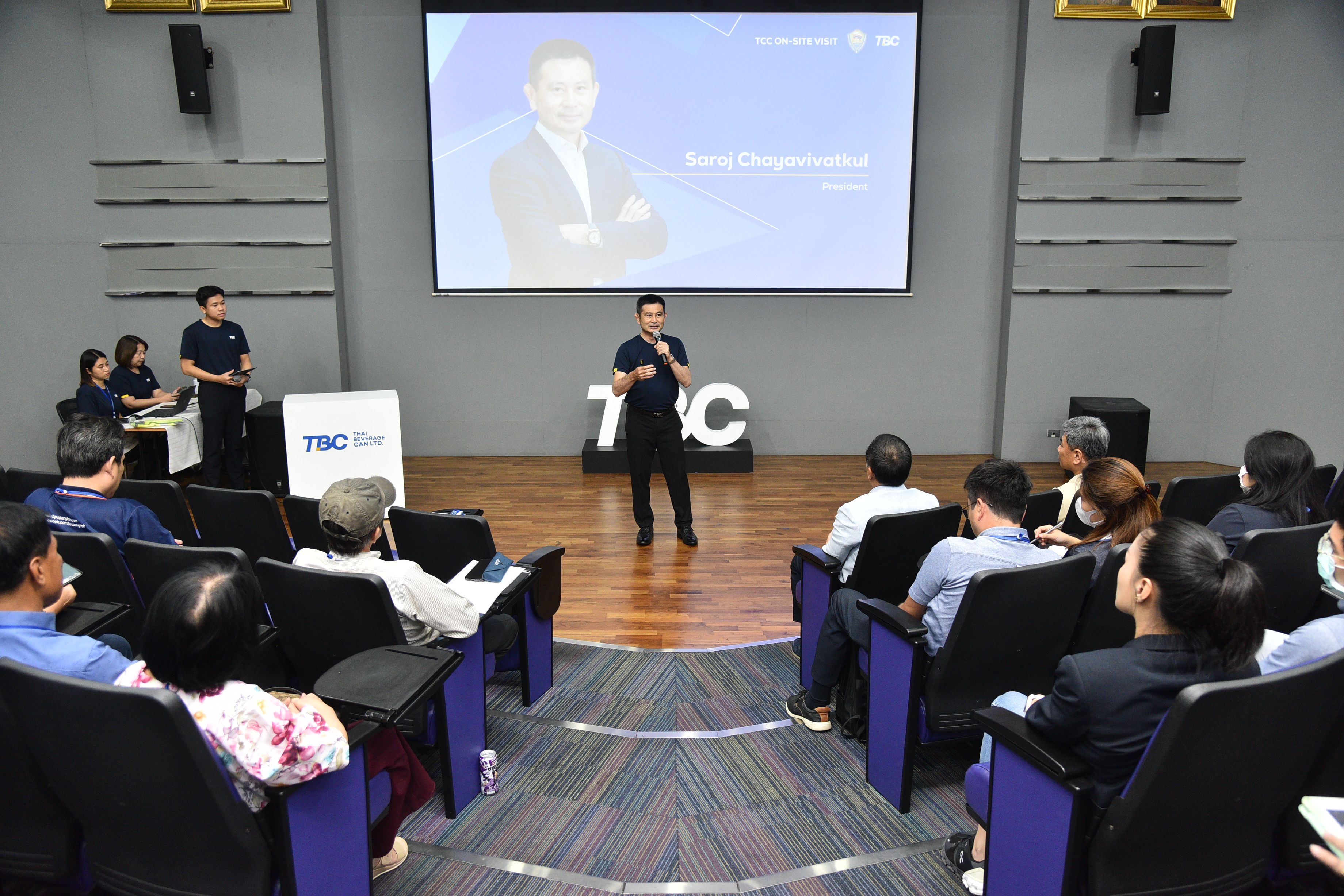 Man speaking on stage in an auditorium with an audience and large presentation screen.