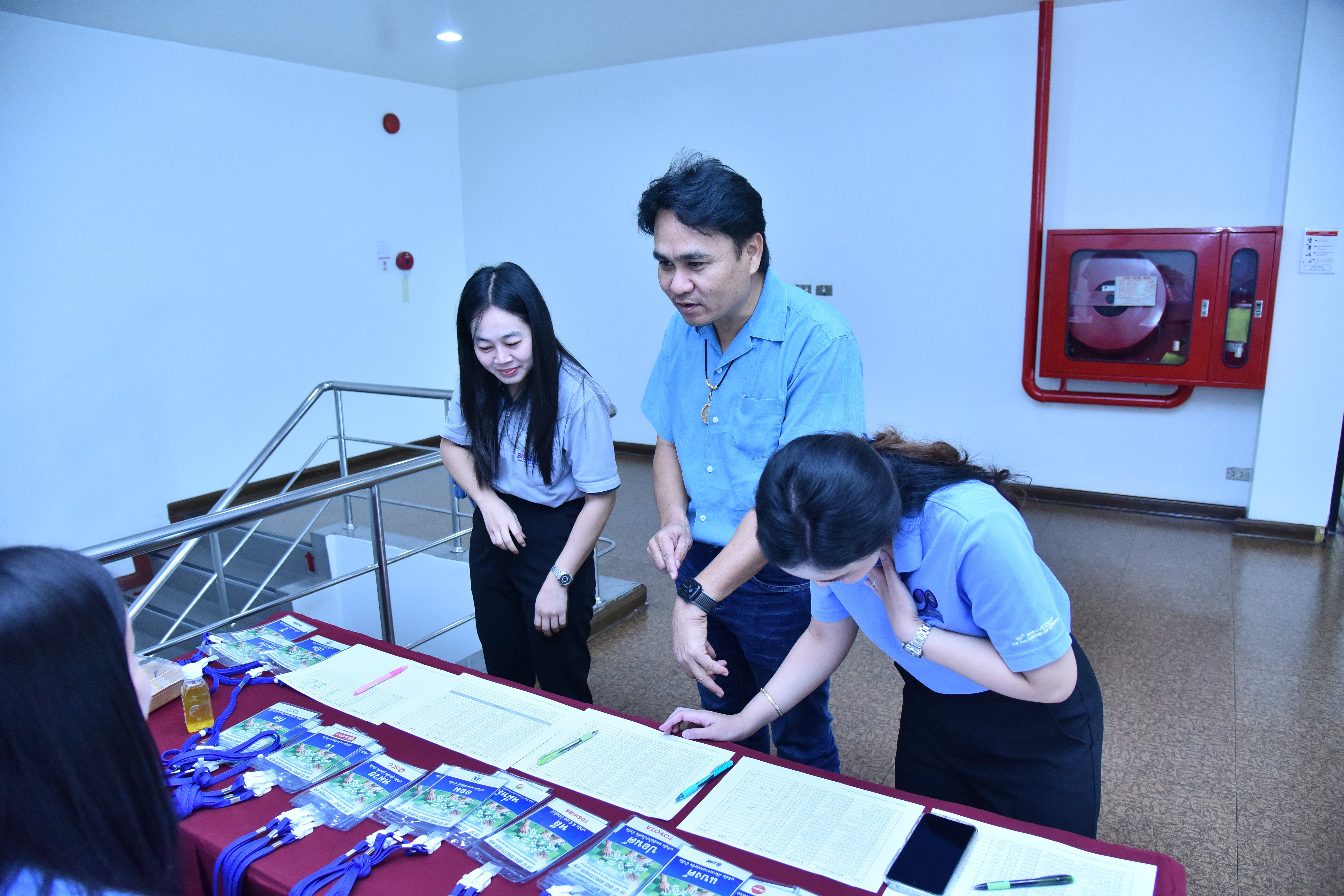 Man and two women review documents and lanyards at a registration table indoors.