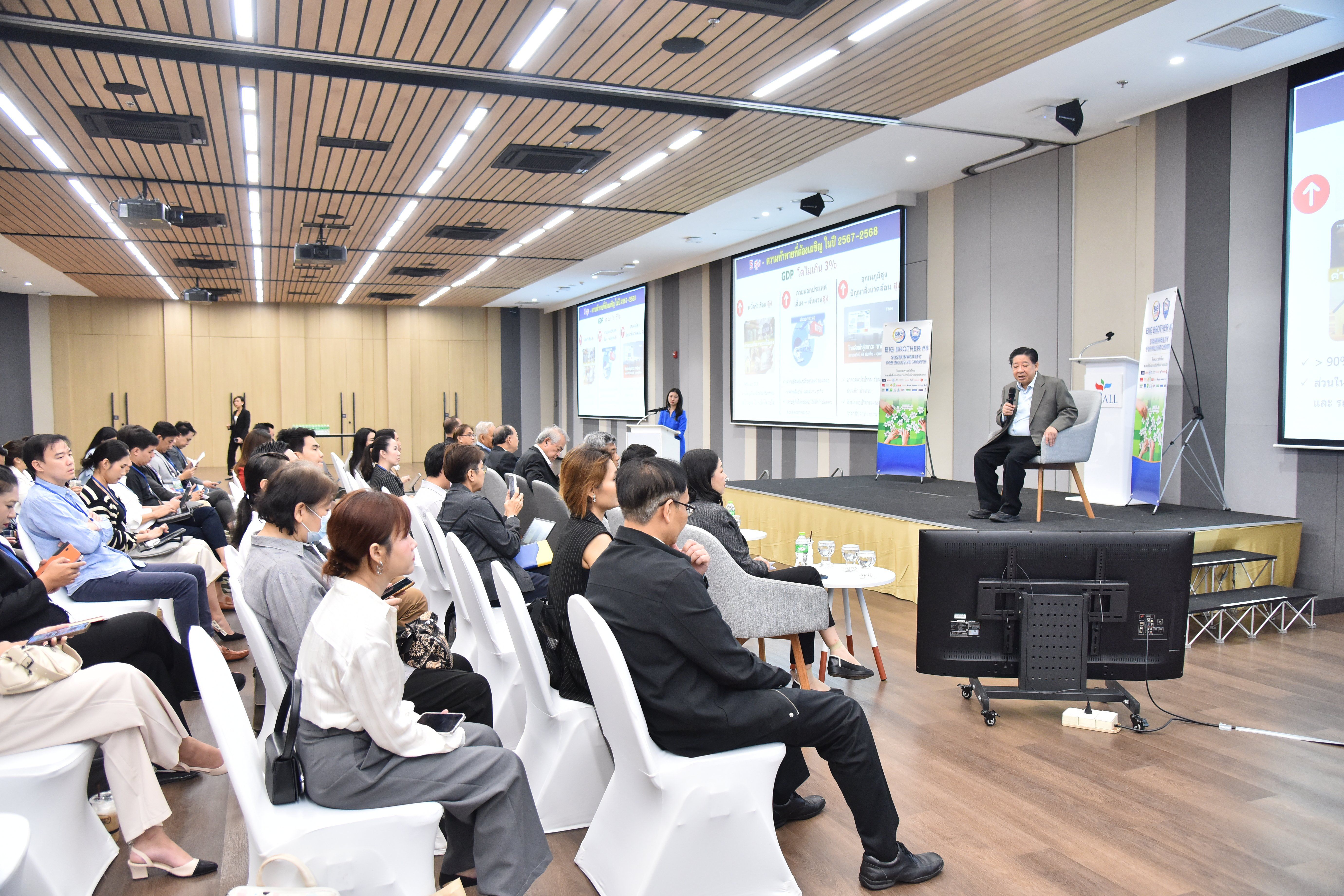 Speaker presents to a seated audience in a bright conference room with large screens.