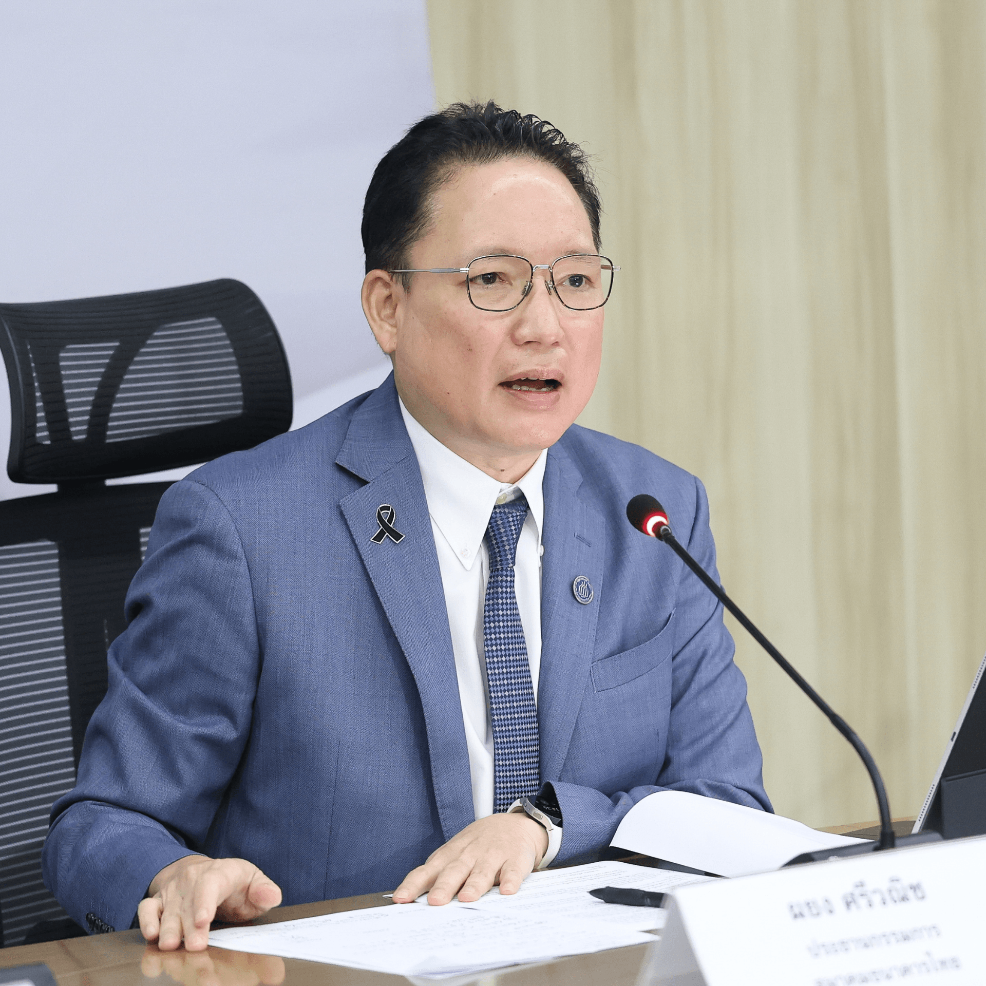 Man in blue suit and glasses speaking at a formal meeting table.
