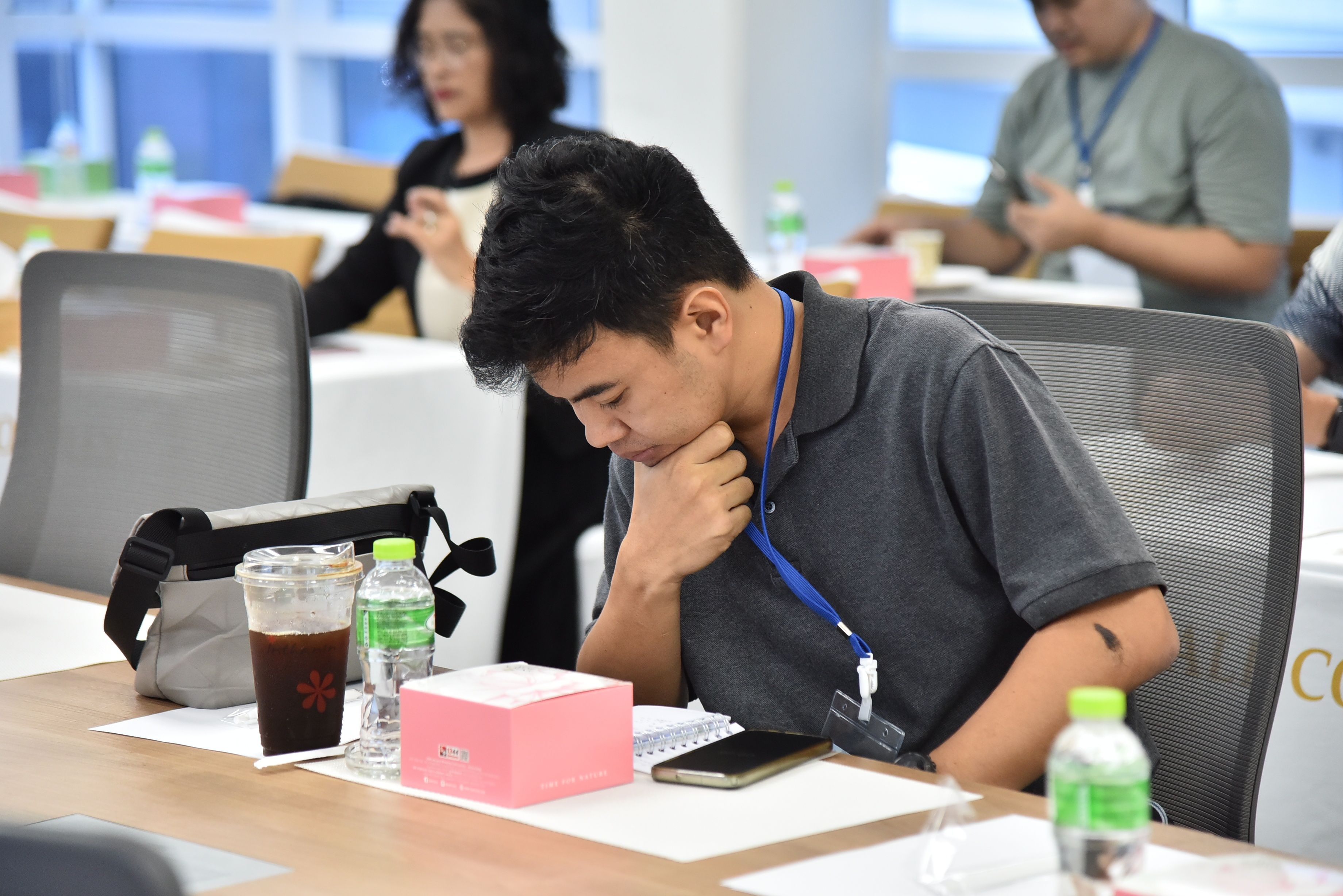Man in grey shirt deeply concentrating at a conference table with phone and drinks.