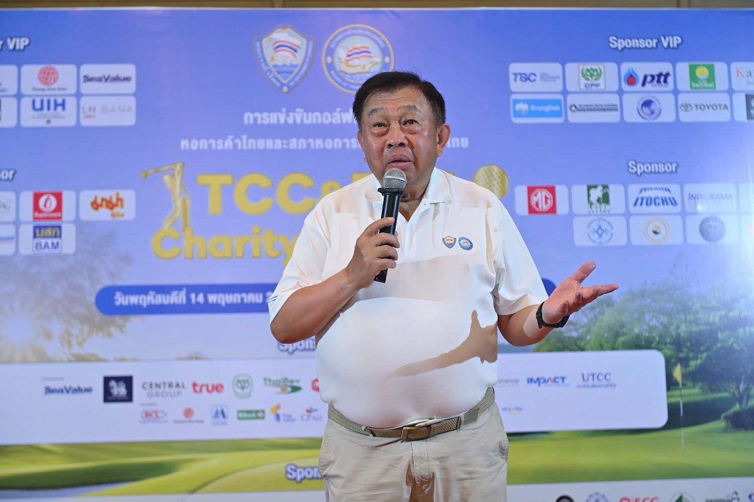 Elderly man speaking into a microphone, gesturing, in front of a 'TCC Charity' banner.