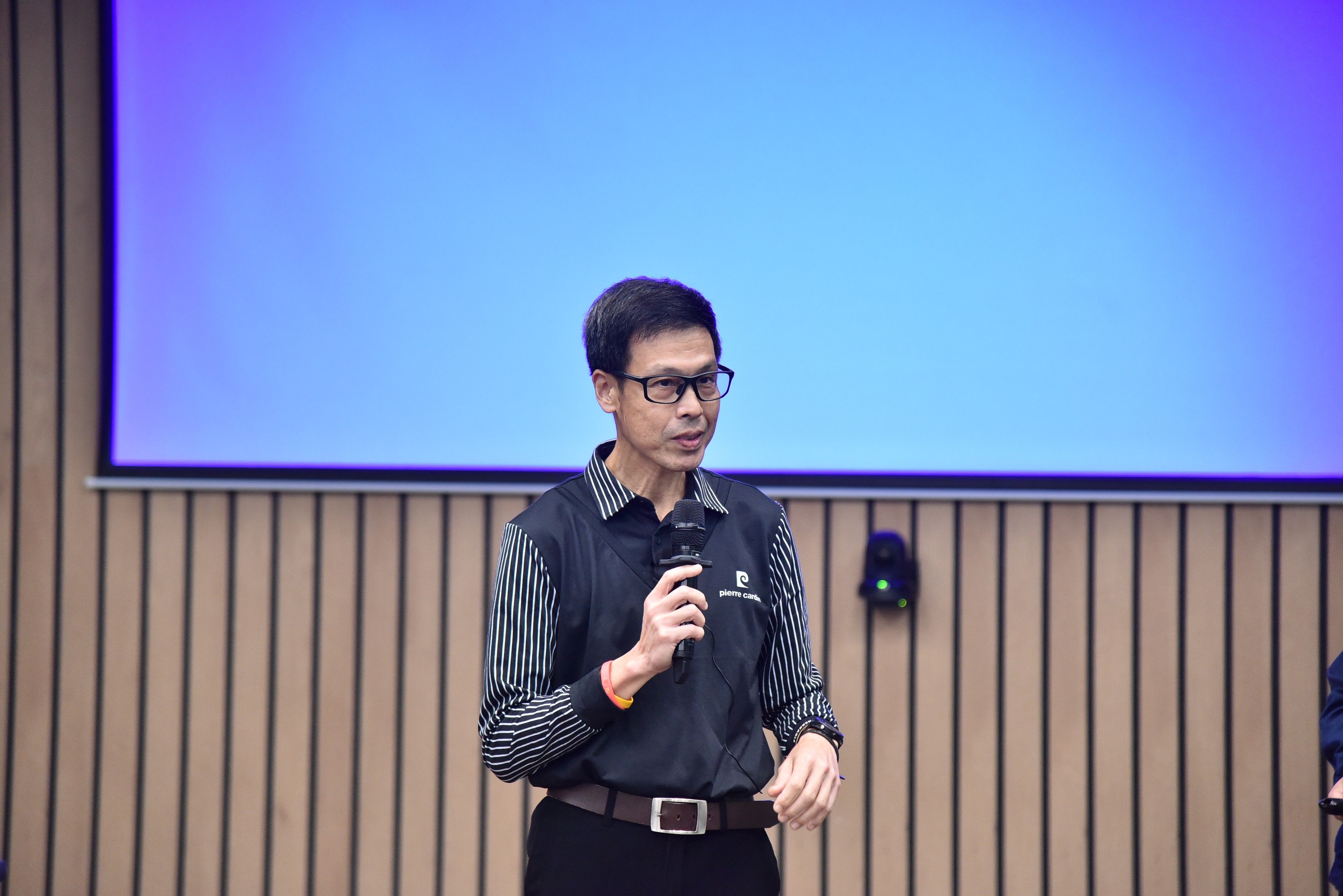 Man in glasses speaks into a microphone on stage before a large blue screen.