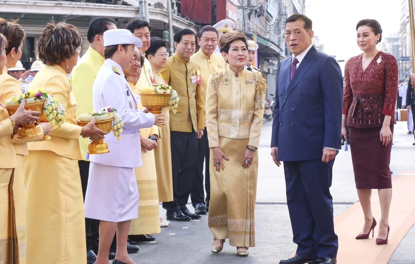 Thai people in traditional dress present offerings in golden bowls to a distinguished couple.