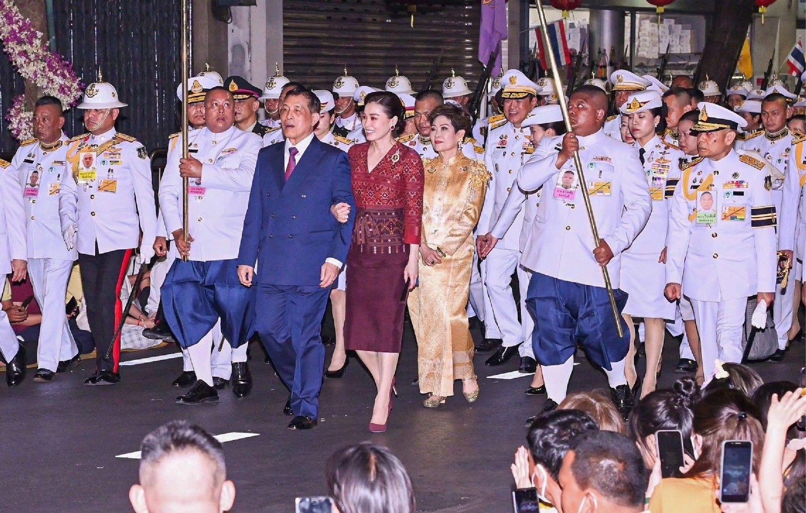 A royal couple and another woman in traditional attire lead a formal procession.