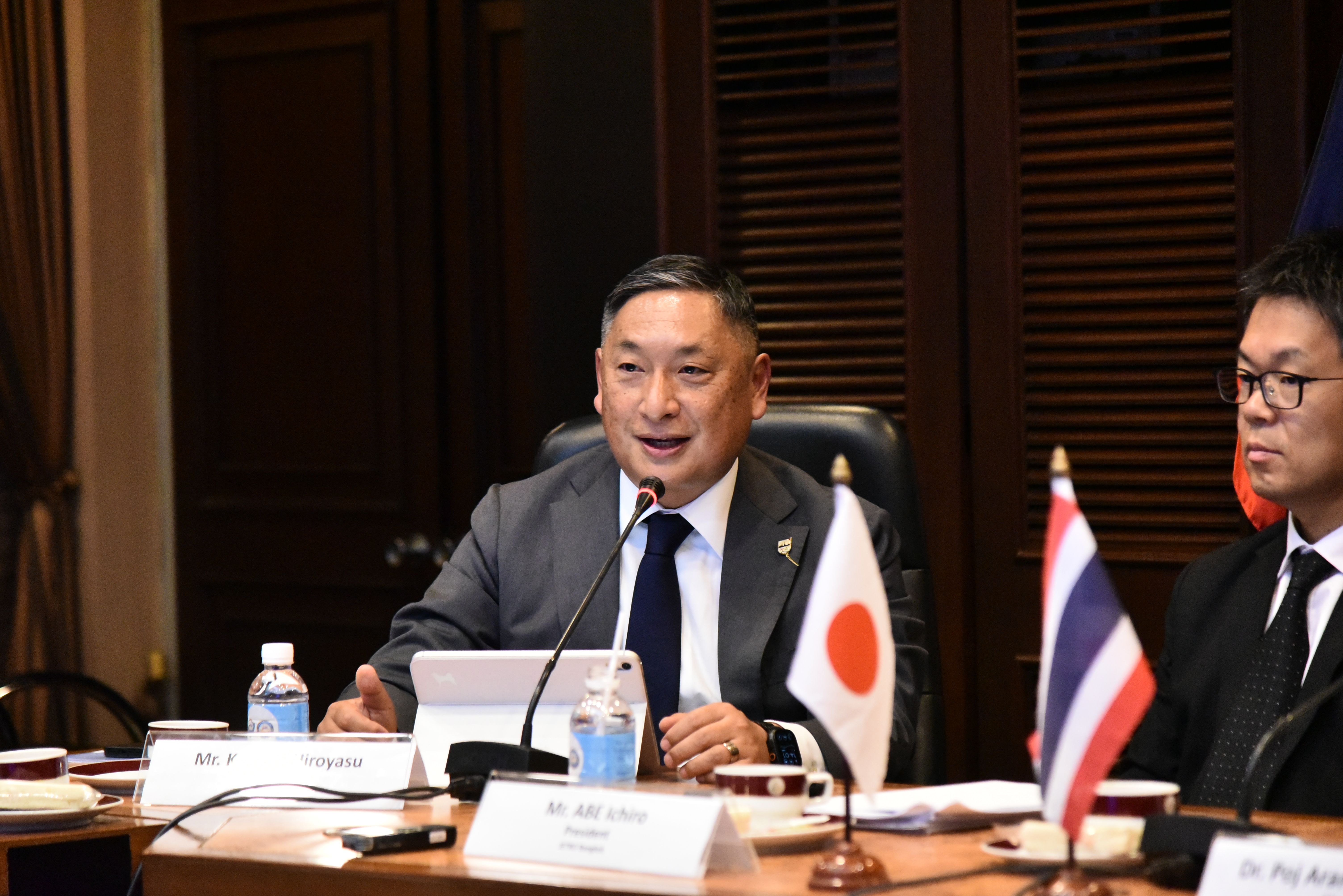 A Japanese man speaks at a formal meeting with Japanese and Thai flags on the table.