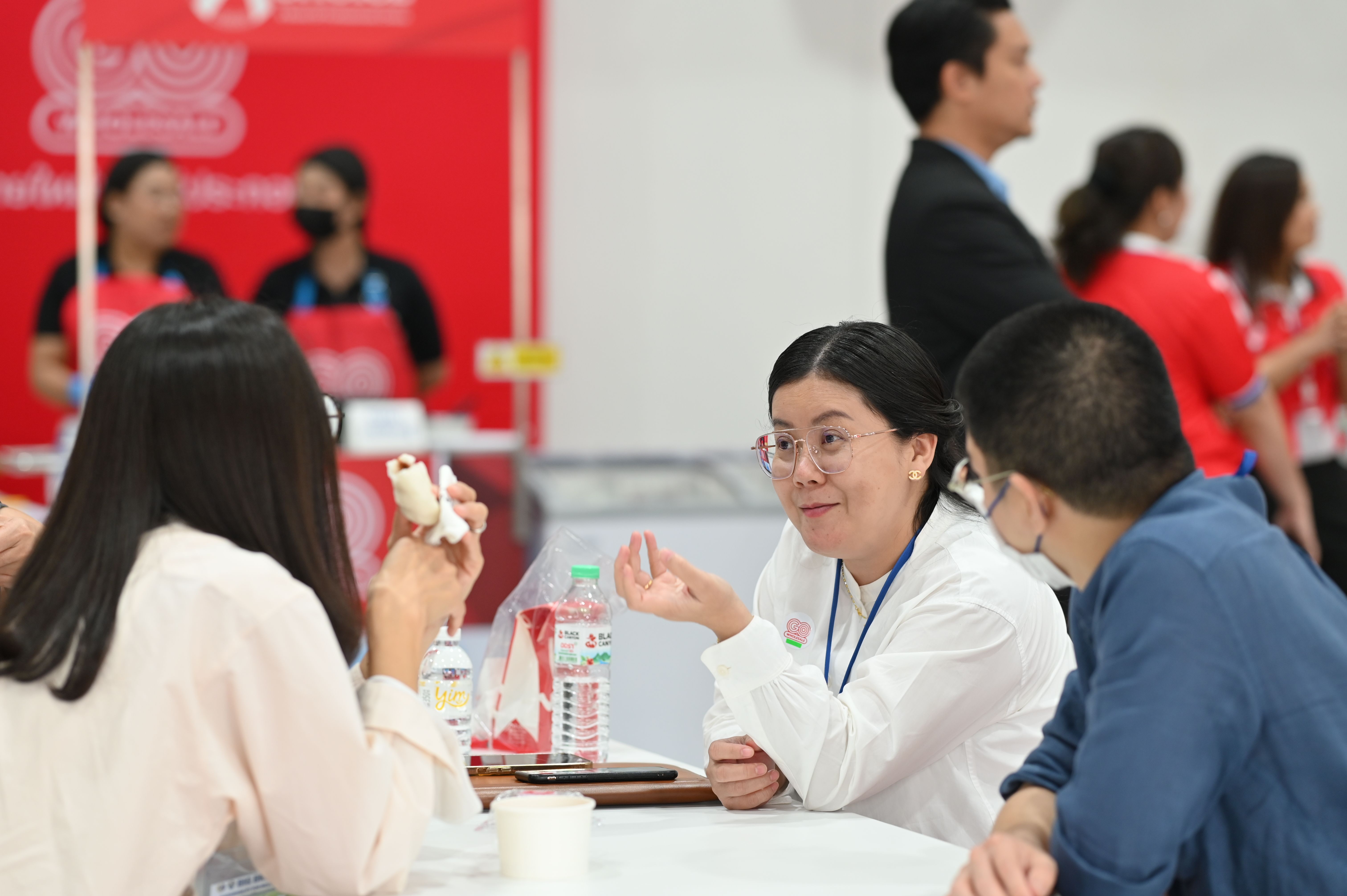 Three people discussing at a table during a busy indoor event with a red booth.