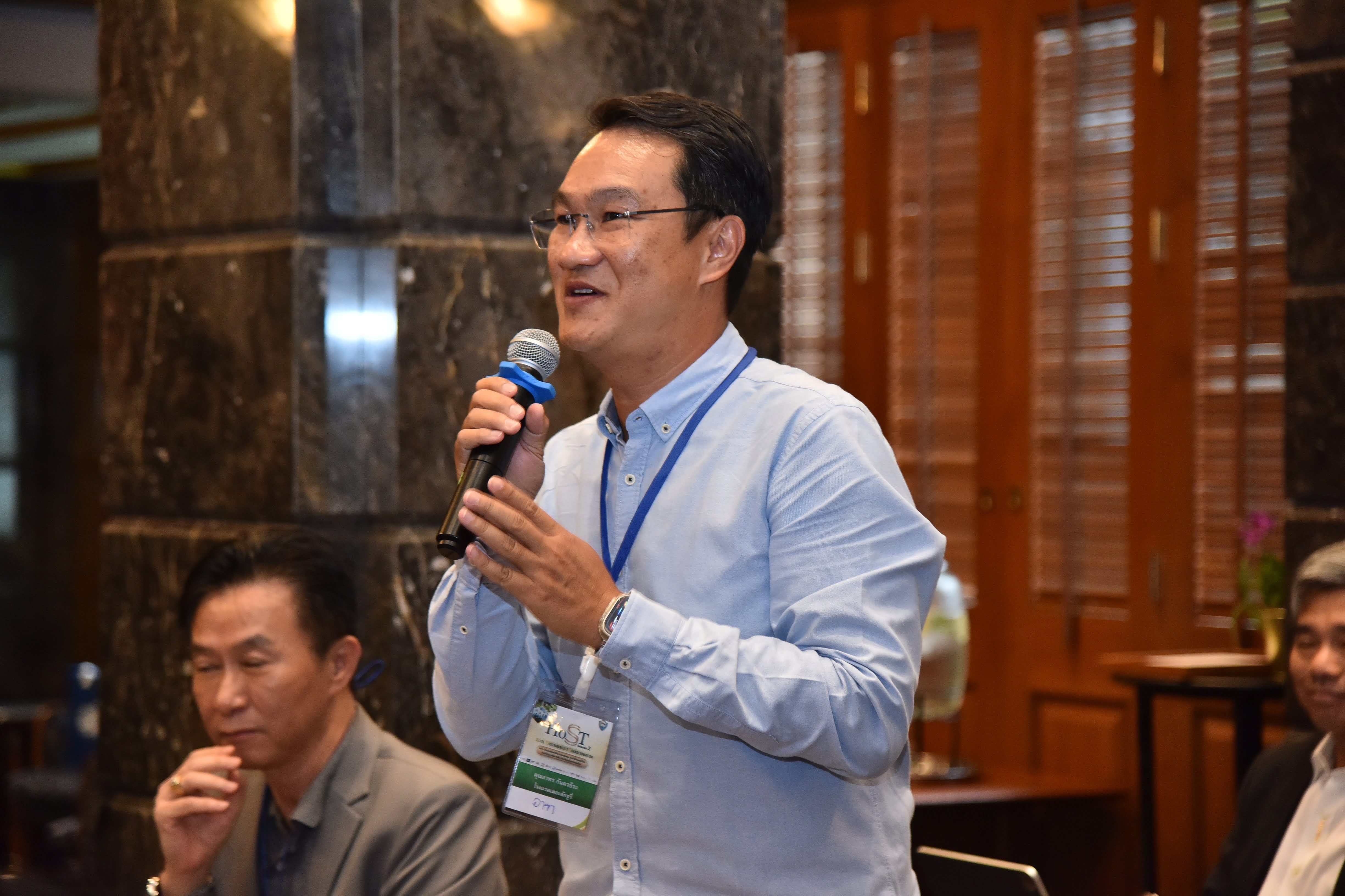 Man in light blue shirt speaks into microphone at an indoor professional event.