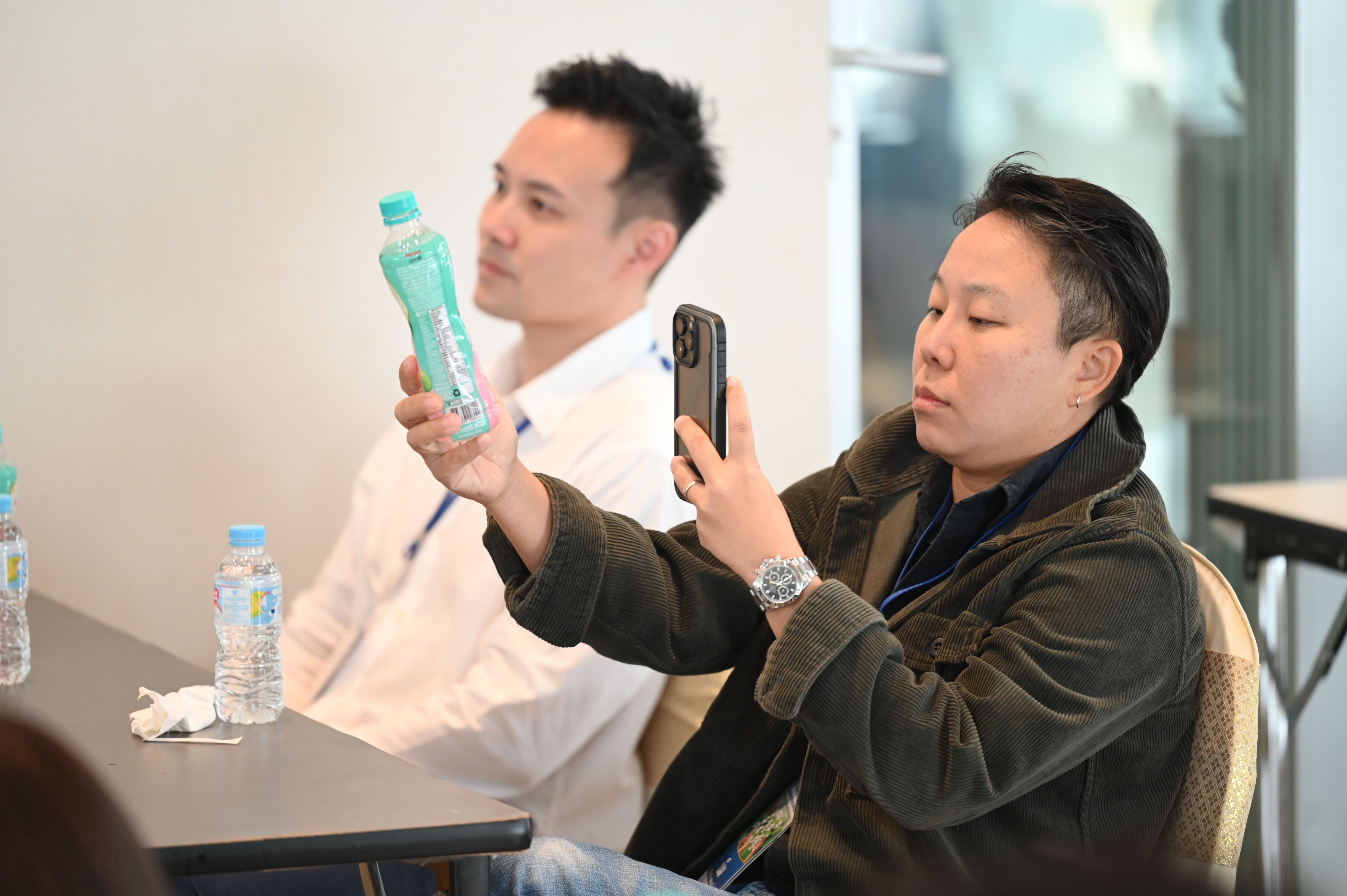 A person uses a smartphone to photograph a light green water bottle held by another.