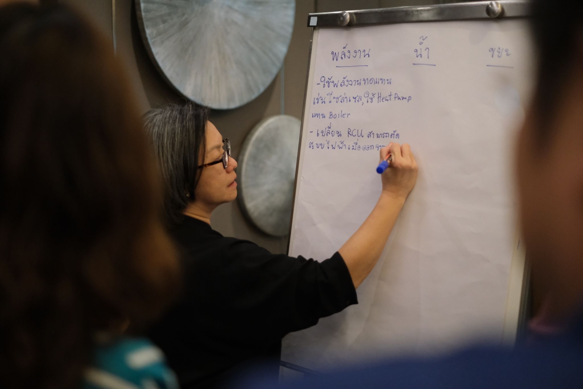 A woman in glasses writes on a whiteboard with a purple marker during a meeting.