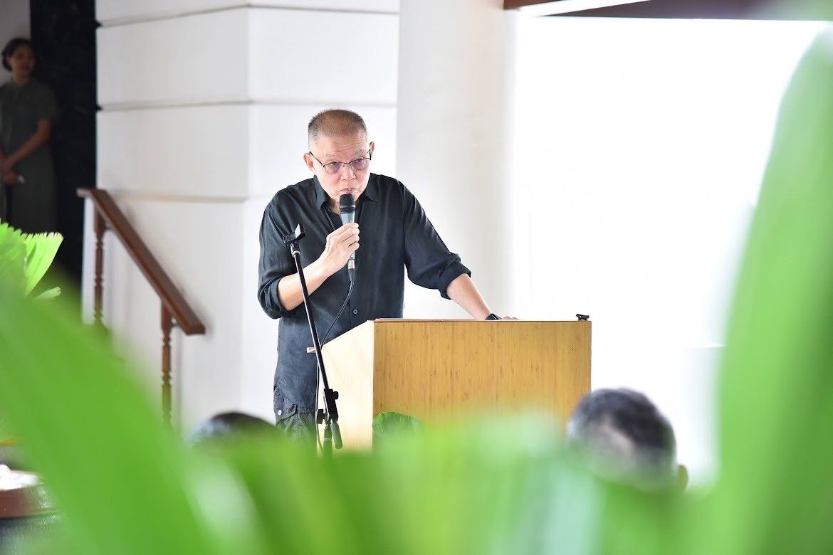 Man in dark shirt speaks into a microphone at a wooden podium during a presentation.