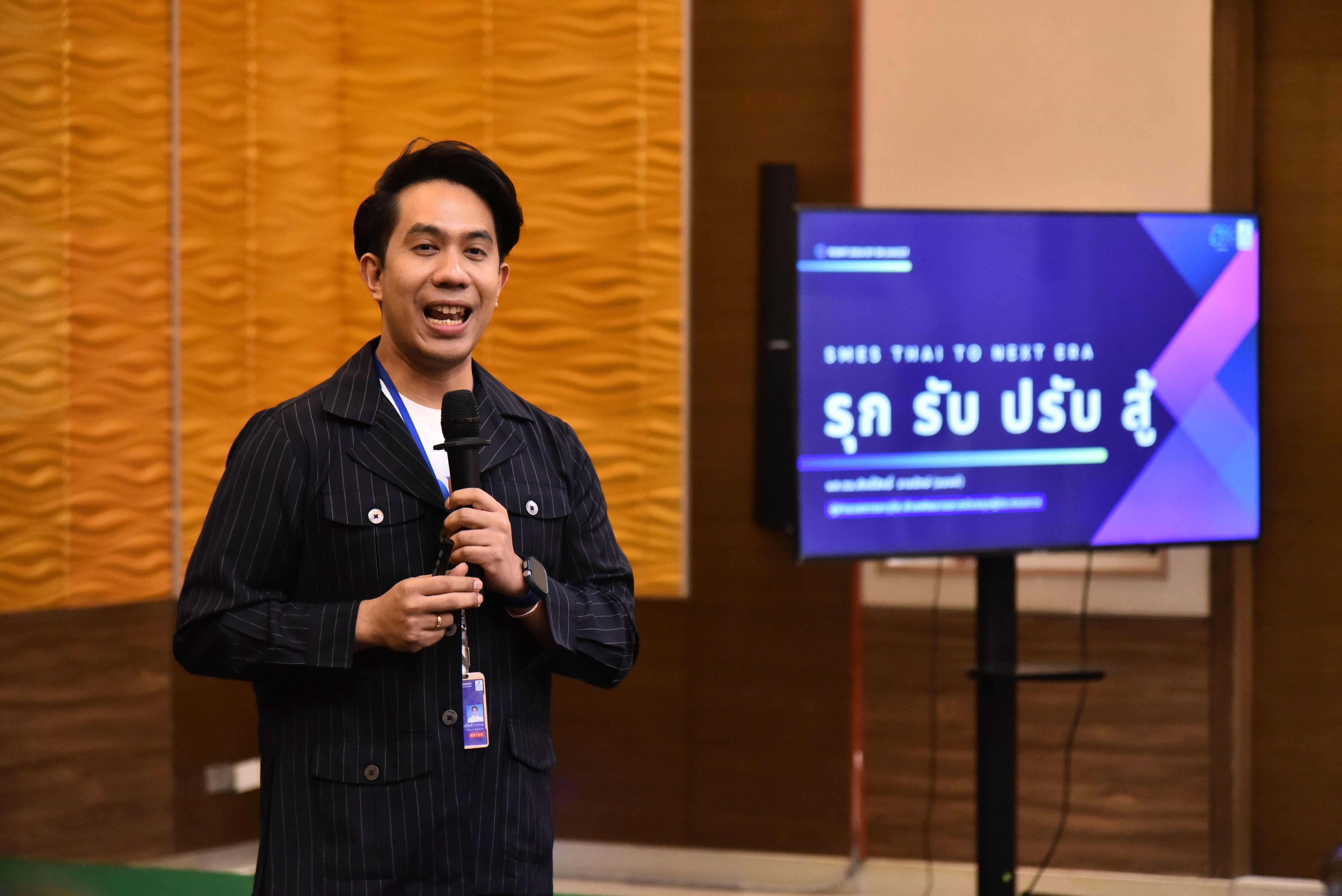 Man speaking into a microphone on stage next to a presentation screen with text.