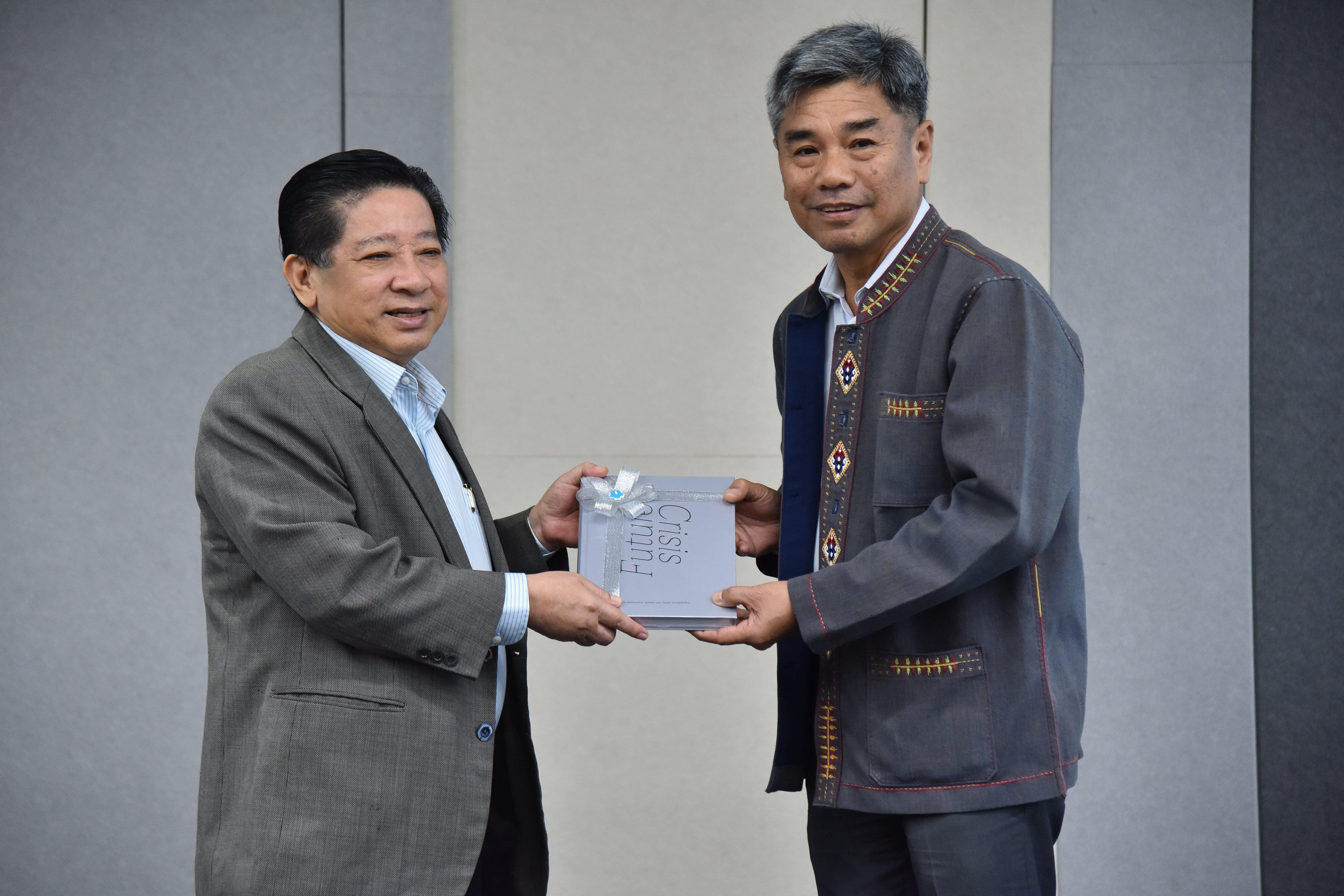 Two Asian men formally exchange a white book or plaque, smiling slightly.
