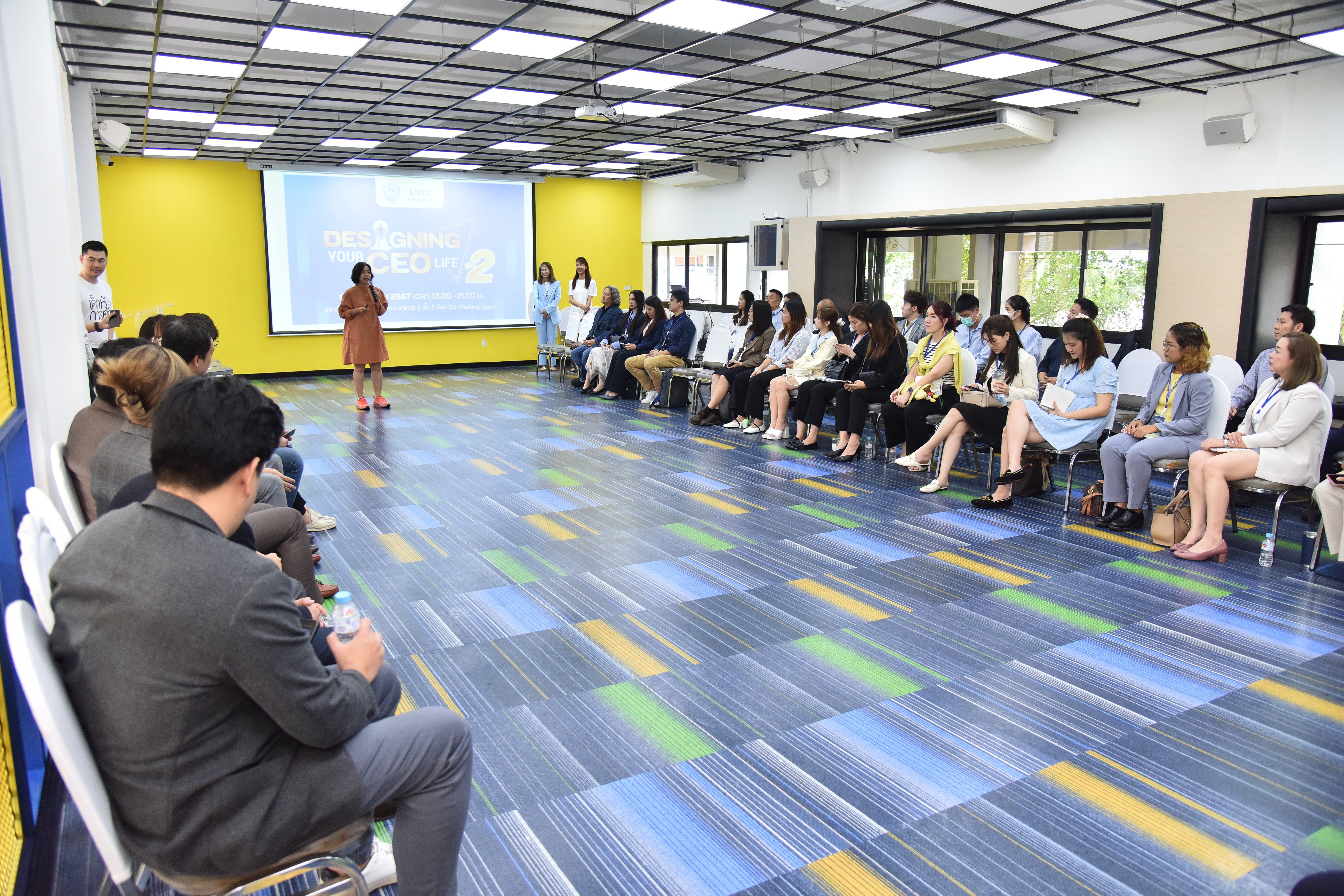 Woman presents to a seated audience in a bright room with colorful floor.
