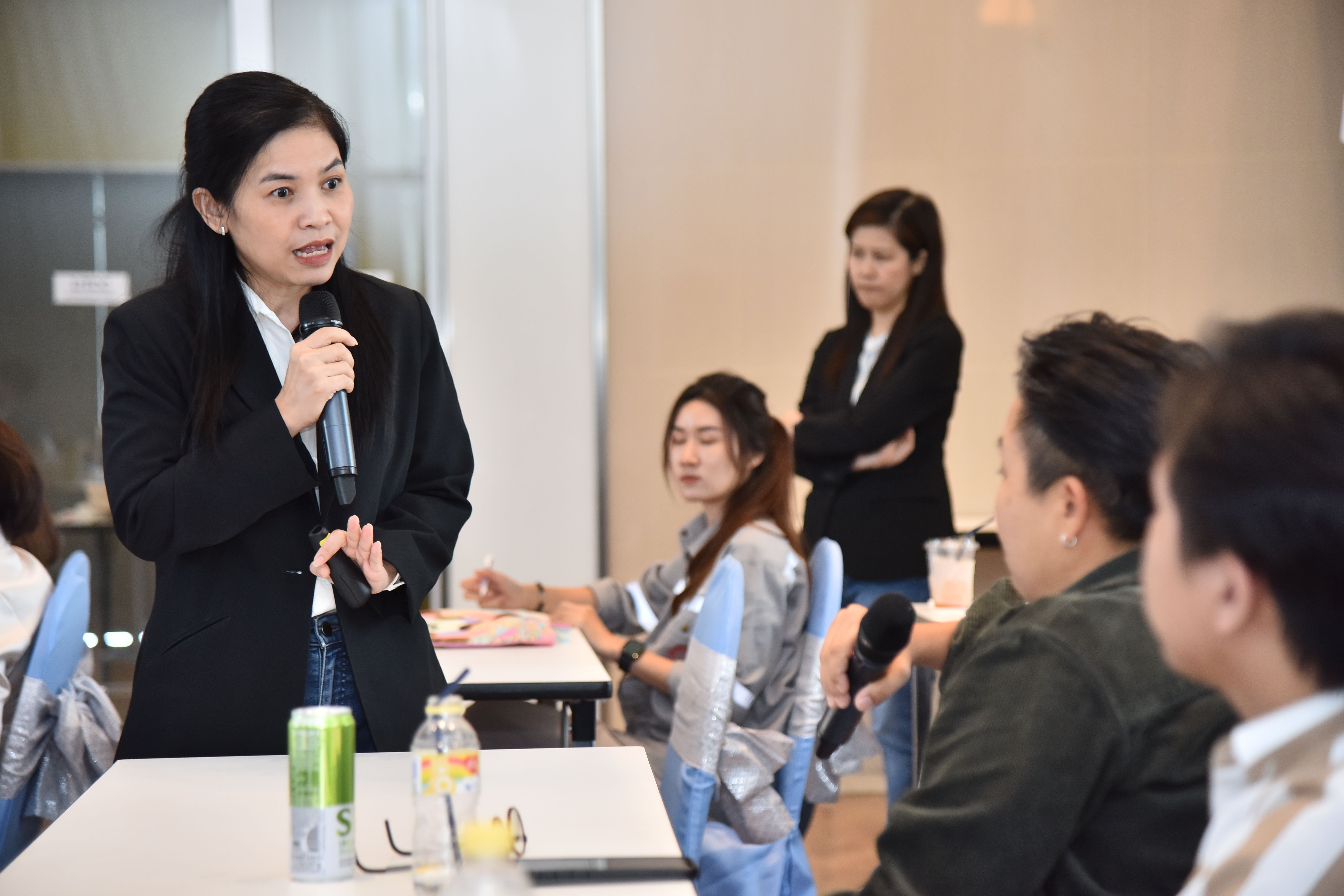 Woman in black suit speaks into microphone during an indoor presentation to seated attendees.