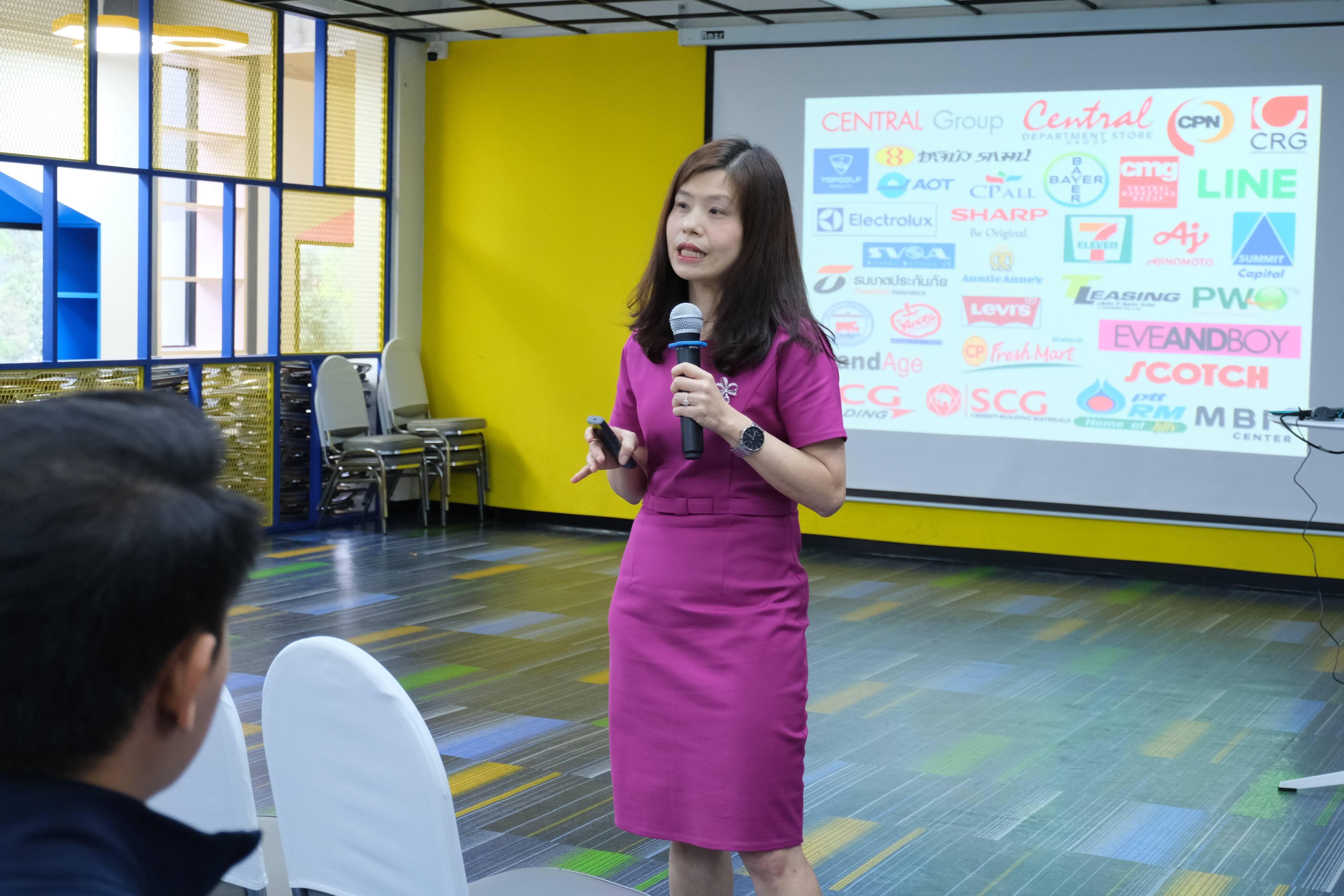 Woman in pink dress speaks, presenting company logos on a screen to an audience.
