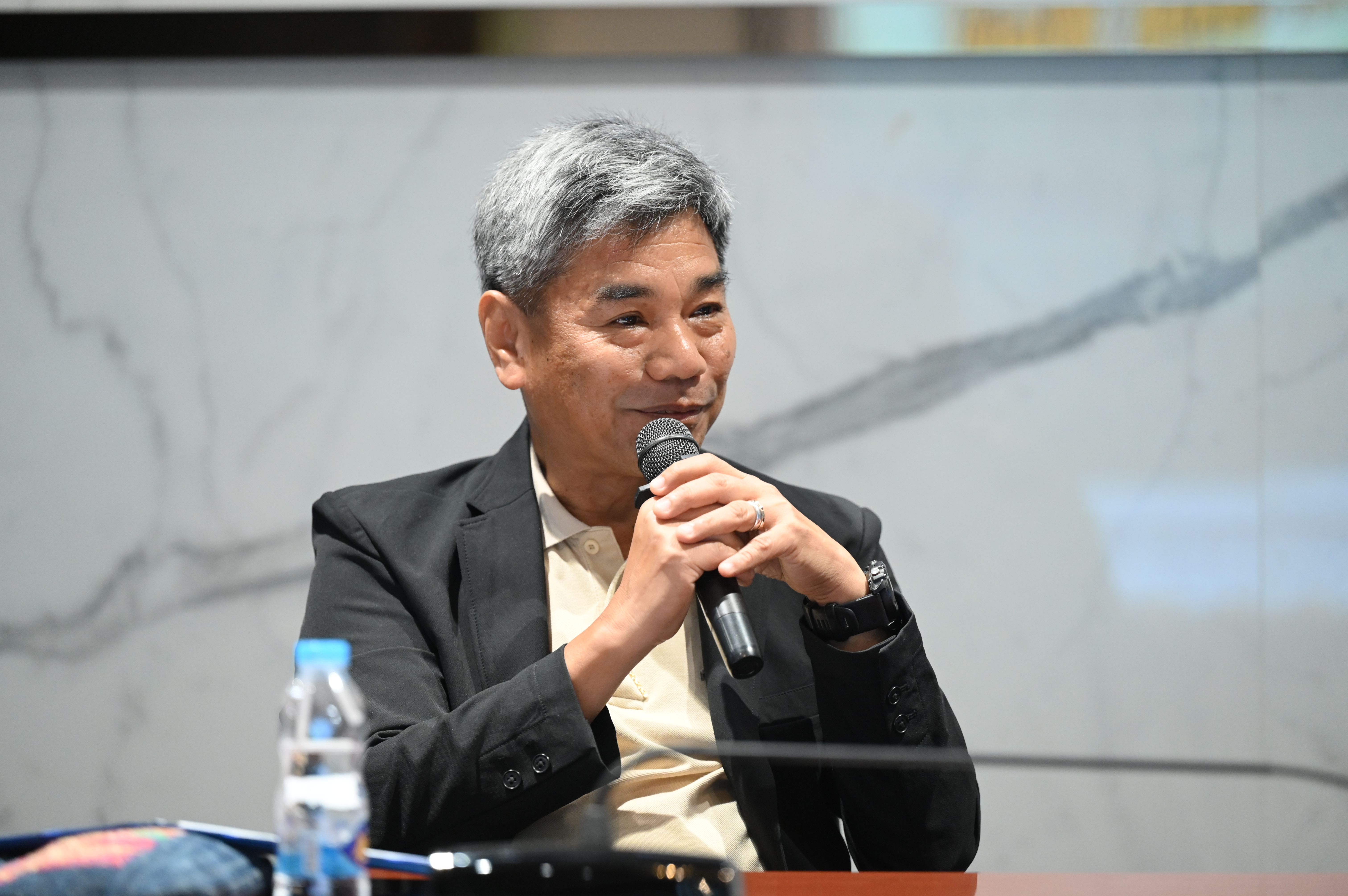 Man with grey hair and black blazer speaks into a microphone at a presentation.