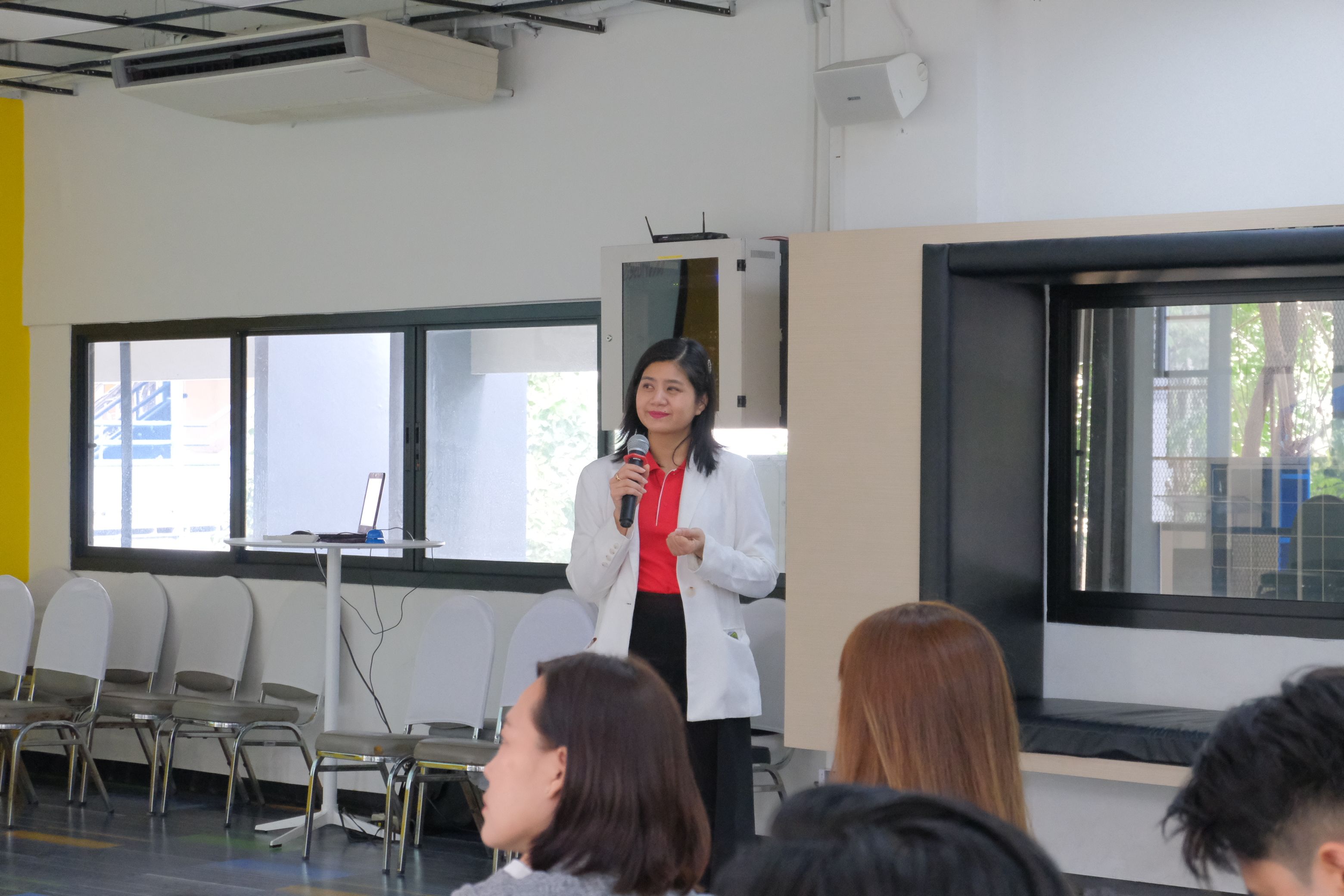 Woman in white blazer speaks into a microphone at an indoor presentation.